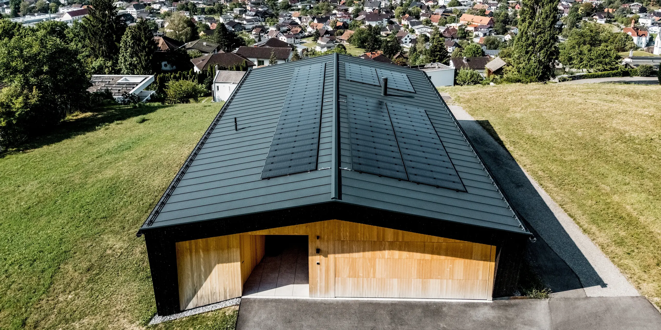 Diese spektakuläre Vogelperspektive zeigt das moderne Einfamilienhaus in Röthis mit einem PREFALZ Satteldach in der Farbe P.10 Anthrazit und den harmonisch integrierten Solarmodulen PREFALZ. Die langlebige und witterungsbeständige Aluminiumdeckung bildet einen spannenden Kontrast zu den warmen Holzdetails. Abgerundet wird die Kulisse durch die beeindruckende Bergkulisse des Vorarlberger Rheintals.