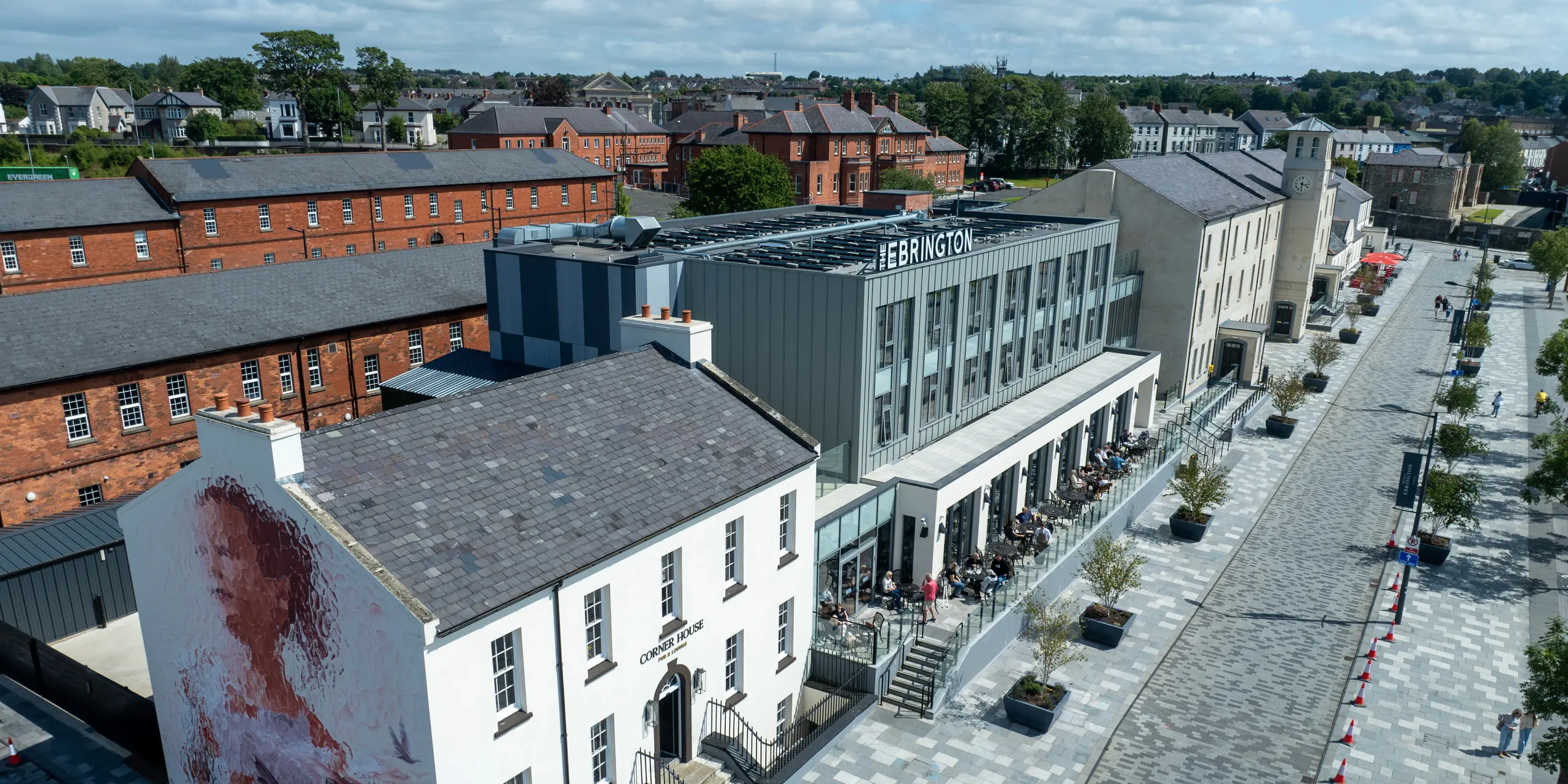 Das Ebrington Hotel im Zentrum von Londonderry, Nordirland, beeindruckt mit seiner modernen PREFALZ-Blechfassade in P.10 Hellgrau und P.10 Zinkgrau. Diese stilvolle und hochwertige Verkleidung von PREFA verleiht dem Gebäude eine elegante Optik und robusten Schutz. Das Bild zeigt auch die Umgebung mit historischen Gebäuden, die einen interessanten Kontrast zur zeitgenössischen Architektur des Hotels bieten. Die langlebige Aluminiumfassade sorgt dauerhaft für eine ästhetische Außenansicht.