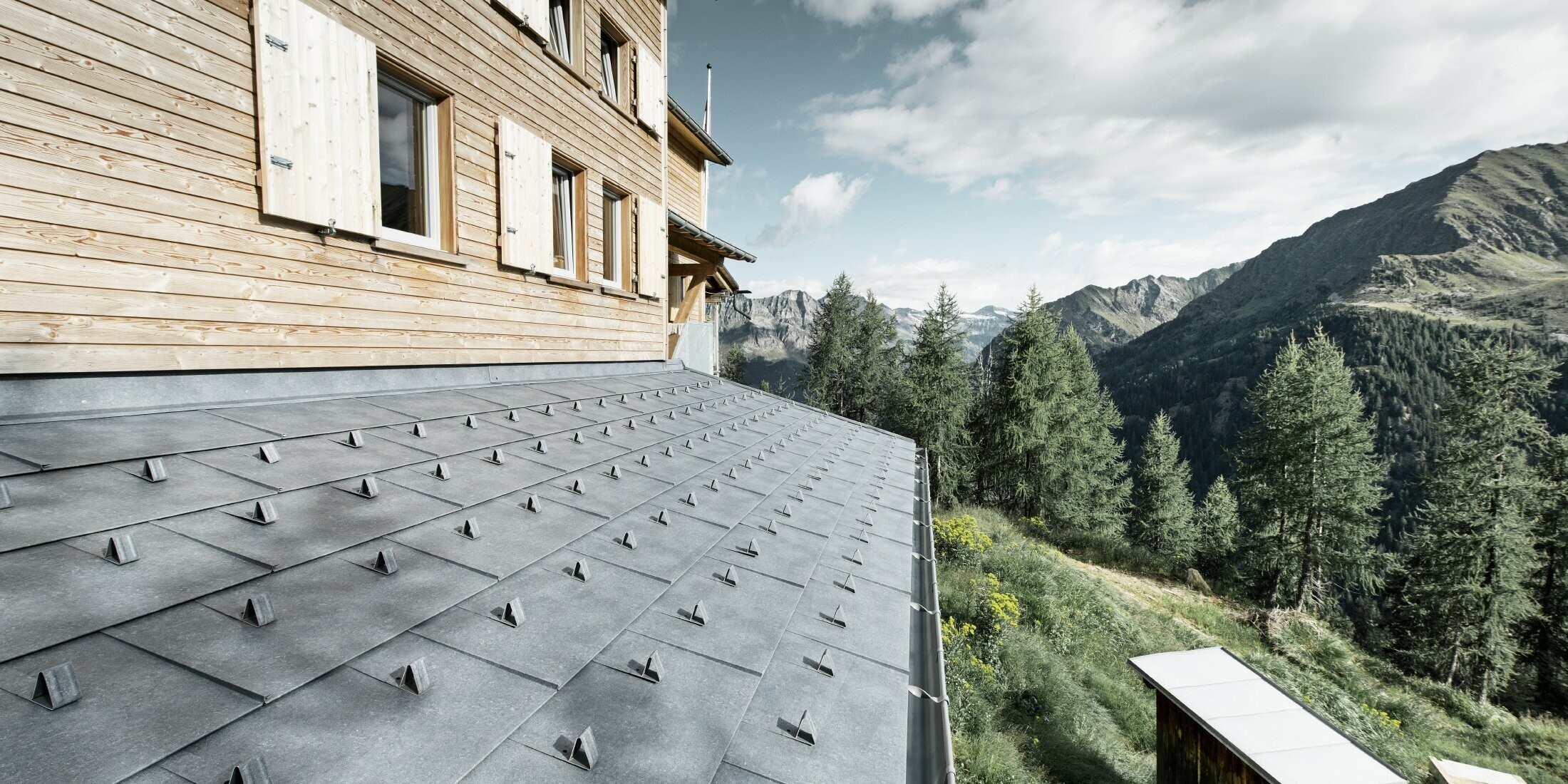 Gros plan sur l’avant-toit d'un refuge en bois. L'avant-toit est recouvert de panneaux d’aluminium FX.12 PREFA couleur gris pierre avec arrêts de neige. La gouttière est maintenue par des brides de renfort.