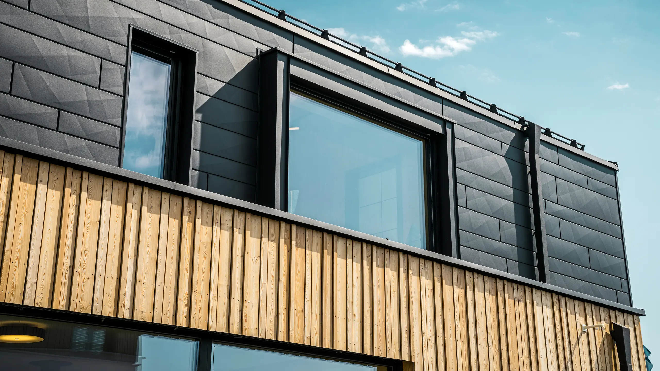 Close-up van de bovenverdieping van het modelhuis Autark in het prefabhuiscentrum Blaue Lagune, Vösendorf (AT). De gevel bestaat uit PREFA Siding.X aluminium panelen in P.10 zwart, die door hun speciale optiek met verschillende kantingen opvallen. Daaronder bevindt zich een stijlvolle houten bekleding, die een harmonieus contrast vormt met het donkere aluminium. Grote ramen laten veel licht binnen in het gebouw en benadrukken het moderne design.