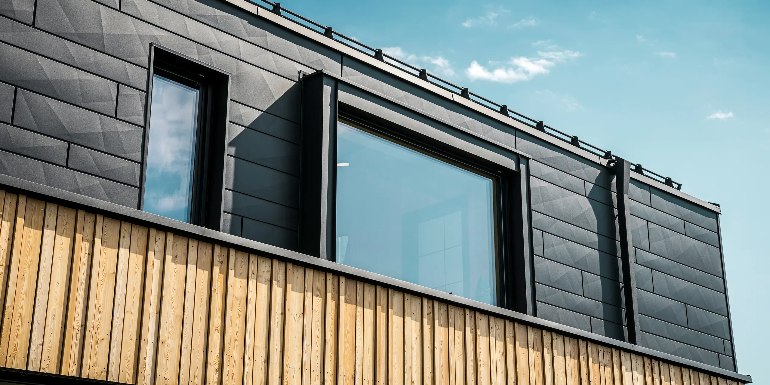 Close-up van de bovenverdieping van het modelhuis Autark in het prefabhuiscentrum Blaue Lagune, Vösendorf (AT). De gevel bestaat uit PREFA Siding.X aluminium panelen in P.10 zwart, die door hun speciale optiek met verschillende kantingen opvallen. Daaronder bevindt zich een stijlvolle houten bekleding, die een harmonieus contrast vormt met het donkere aluminium. Grote ramen laten veel licht binnen in het gebouw en benadrukken het moderne design.