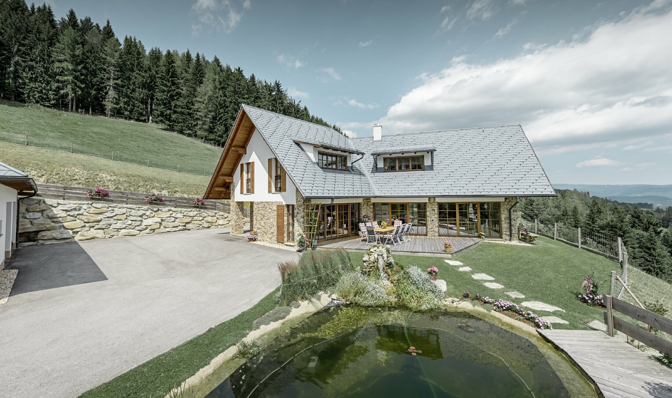 Das Einfamilienhaus in Hanglage, in L-Form gebaut, hat ein Satteldach mit zwei Schleppgauben. Das Dach wurde mit der PREFA Dachschindel in Steingrau eingedeckt. Im Vordergrund ist ein Teich angelegt und im Hintergrund erkennt man einen Wald.