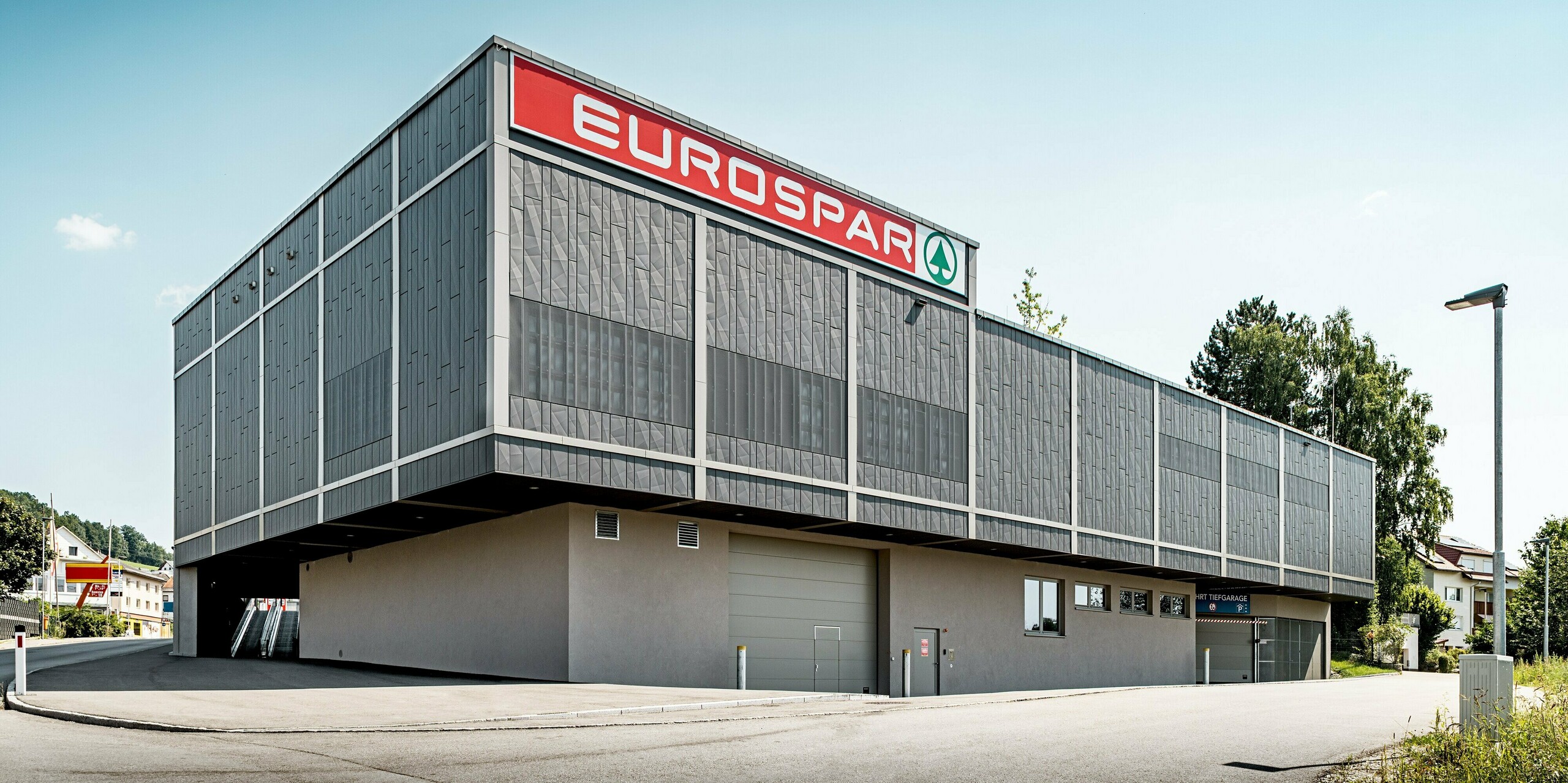 Die Rückseite des Eurospars in Rohrbach mit Tiefgaragenzufahrt. Die moderne Aluminiumfassade von PREFA besteht aus anthrazitfarbenen Siding.X-Paneelen, die durch ihre präzise, geometrische Struktur eine markante und moderne Optik erzeugen. Besonders auffällig sind die perforierten Paneele im oberen Bereich. Sie bieten eine raffinierte Textur und sorgen gleichzeitig für eine gezielte Lichtdurchlässigkeit. Diese Fassadengestaltung verleiht dem Gebäude eine langlebige, pflegeleichte und ästhetisch ansprechende Hülle, die sich perfekt in die Umgebung einfügt.