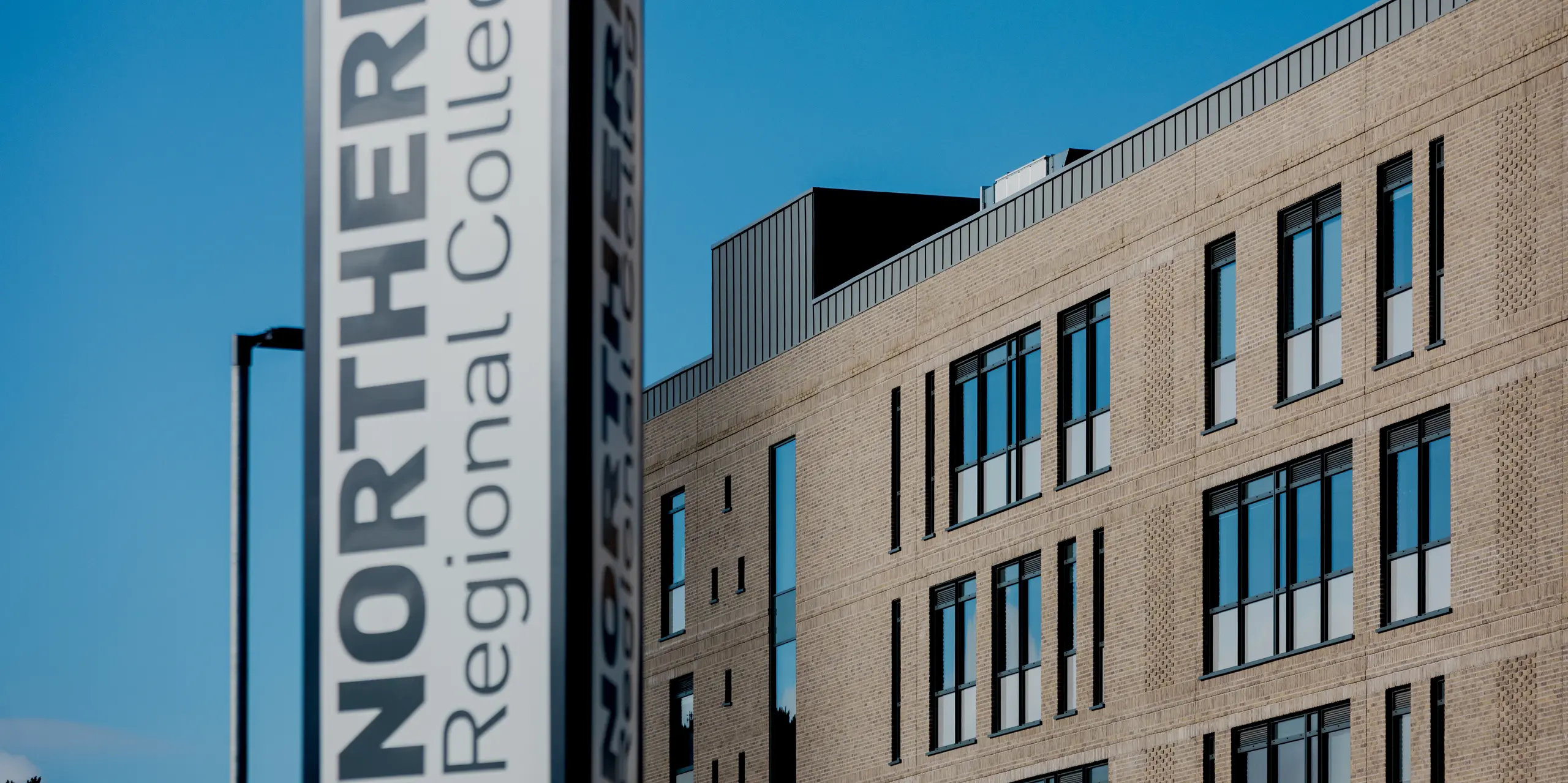 Au premier plan se dresse la stèle verticale portant l'inscription « Northern Regional College », derrière laquelle se dévoile, sous un ciel d'un bleu limpide, la façade en briques du campus de Ballymena. Le long de la toiture, les bacs à joints debout en aluminium PREFALZ, gris quartz, créent une finition horizontale et précise. Les fenêtres étroites structurent la façade avec rythme et soulignent la linéarité de l'architecture. L'aluminium PREFA confère au bâtiment scolaire une allure durable, nécessitant peu d'entretien et d'une modernité intemporelle.