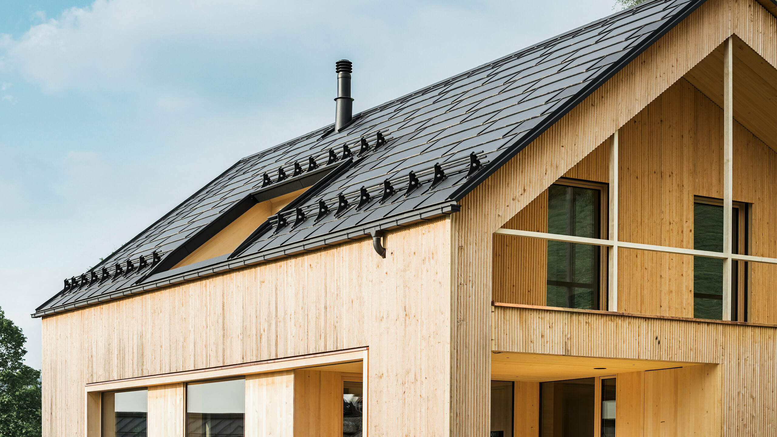 Close-up van de gevel van een milieuvriendelijke woning in Egg, gekenmerkt door PREFA solar-dakpannen in elegant zwart, die harmonieus zijn geïntegreerd in het midden van de PREFA dakpan R.16. De strakke lijnen van het dak contrasteren met de natuurlijke textuur van de houten gevel, terwijl de solarmodules een statement maken voor duurzaam wonen. De subtiele schoorsteen past in het totaalbeeld, dat milieubewust design combineert met landelijke esthetiek. Bovendien beschikt de innovatieve eco-woning over twee balkons die uitzicht bieden op het prachtige berglandschap van Egg in Vorarlberg.
