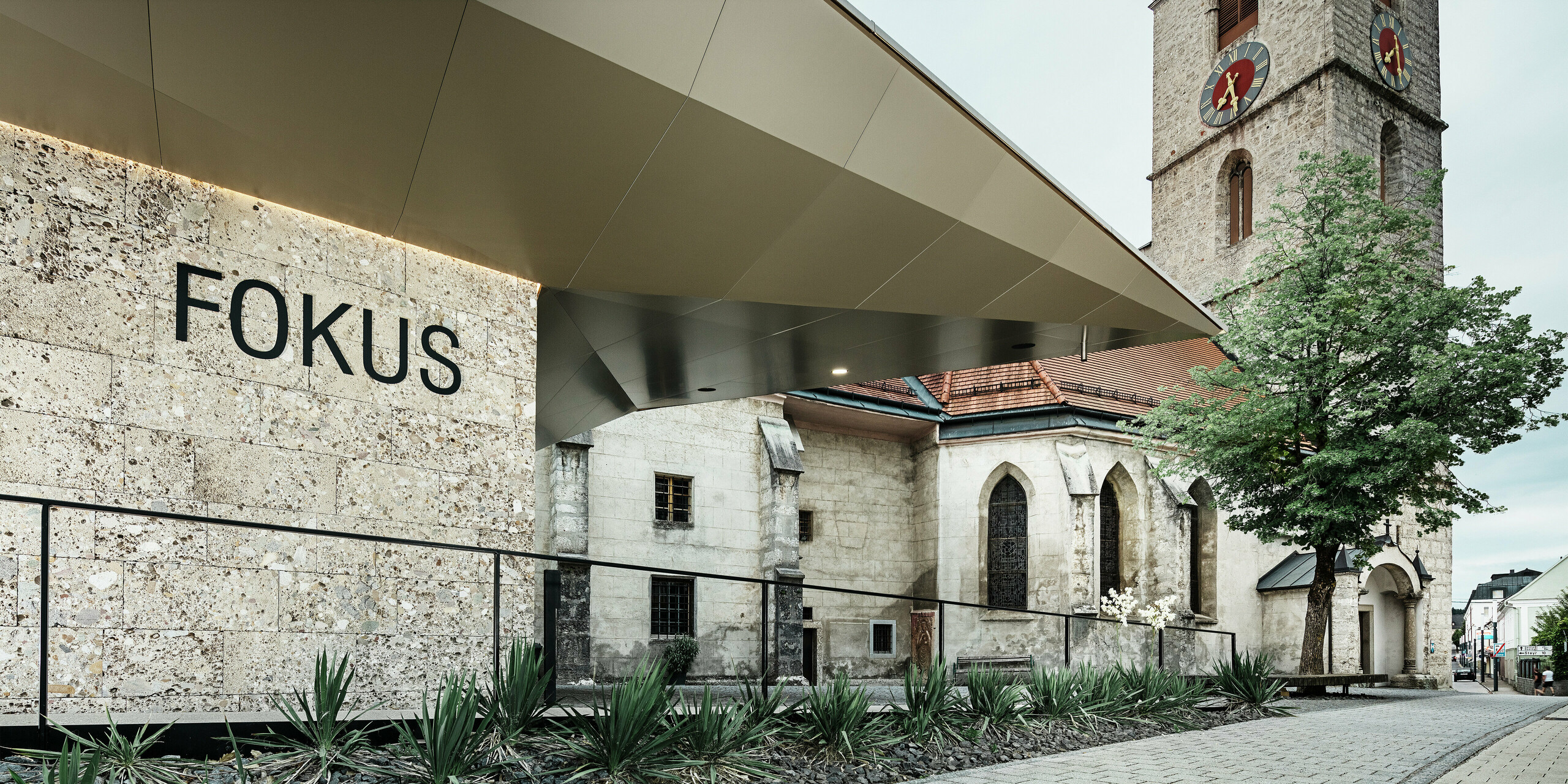 Die klare, reduzierte Geometrie des neuen Baukörpers bildet einen spannenden Kontrast zur historischen Kirche. Die glatten und langlebigen PREFA Aluminium Verbundplatten fügen sich elegant ein und sorgen für eine hochwertige, moderne und wartungsarme Fassadenlösung am Pfarrzentrum in Sierning. Die Perspektive lenkt den Blick auf das neue Foyer mit dem Schriftzug „FOKUS“.