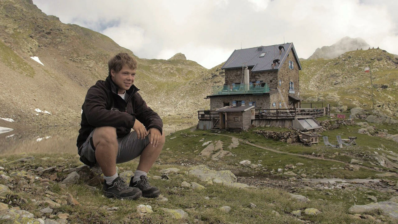 Leerjongen Jakob Schöttl zit op een steen. Op de achtergrond ligt de Flagger Schartenhütte, waarvan het dak werd vernieuwd met PREFA dakpannen.