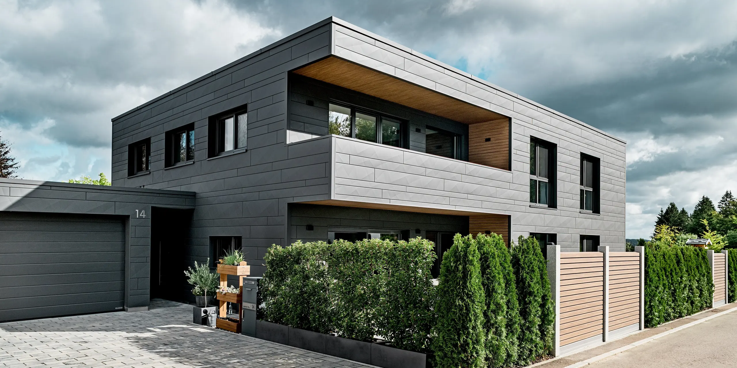 Vue de l'allée d'une maison individuelle moderne à Heidenheim, en Allemagne, revêtue d'une façade en aluminium Siding.X PREFA couleur P.10 gris sombre. Les panneaux aux lignes épurées, de largeurs variables et aux arêtes marquées, créent une façade dynamique. Une haie dense et une clôture en bois apportent un contraste naturel et chaleureux à la surface métallique précise. L'entrée couverte et le grand garage s'intègrent harmonieusement à l'ensemble. La surface en aluminium PREFA, résistante et durable, offre une protection optimale et une architecture moderne et de haute qualité.