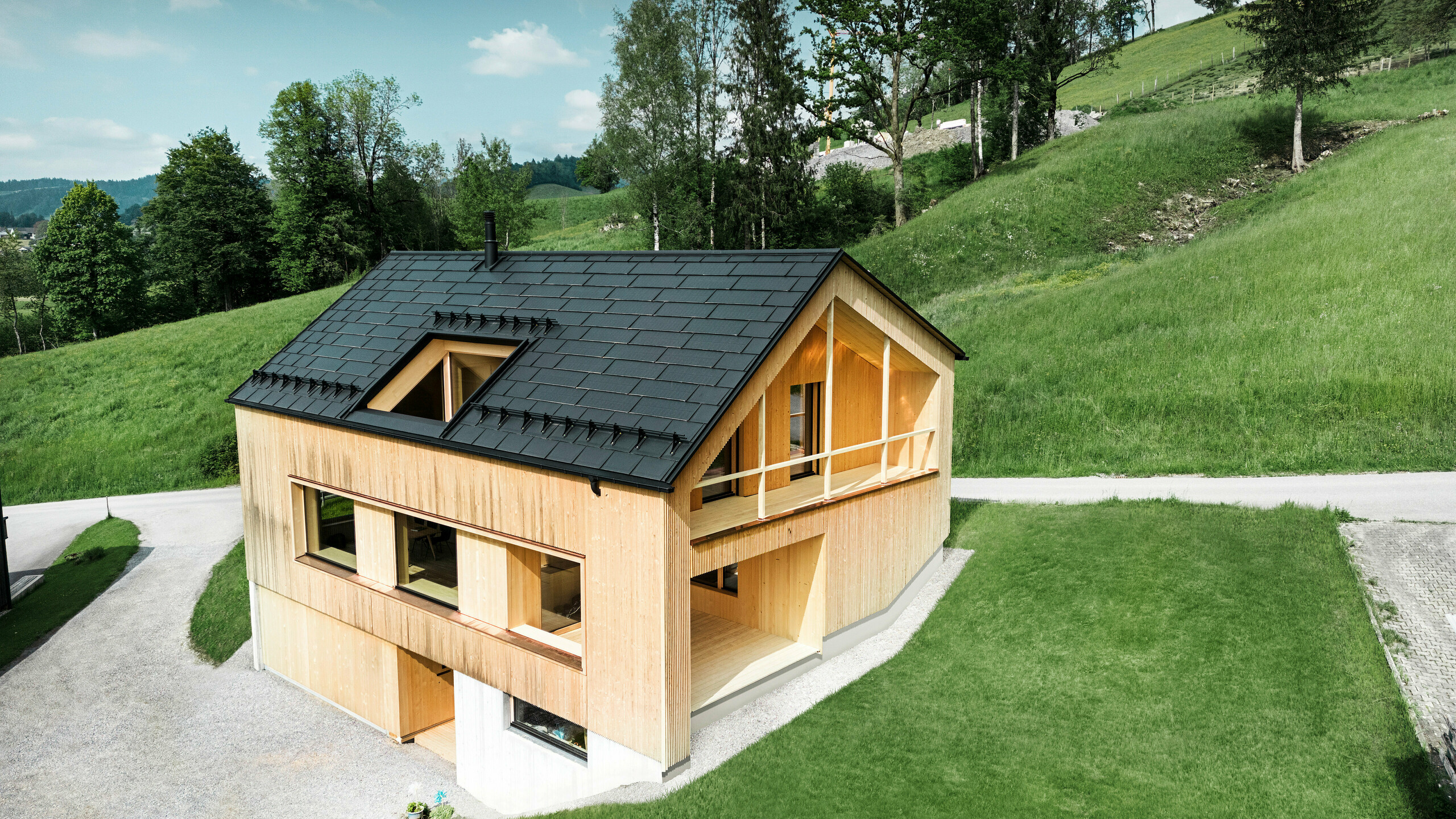 La photo présente une maison écologique pittoresque à Egg, nichée dans les collines verdoyantes, avec un toit solaire PREFA durable. Les R.16 PREFA noirs de haute qualité et les tuiles solaires PREFA s'intègrent harmonieusement dans le paysage rural et soulignent la beauté naturelle des environs. Les tuiles solaires ne sont pas seulement un engagement en faveur des énergies renouvelables, mais aussi un élément de design qui complète avec style la façade en bois moderne et les lignes épurées du bâtiment. Cette maison est un modèle de construction respectueuse de l’environnement et une source d’inspiration pour une architecture tournée vers l’avenir.