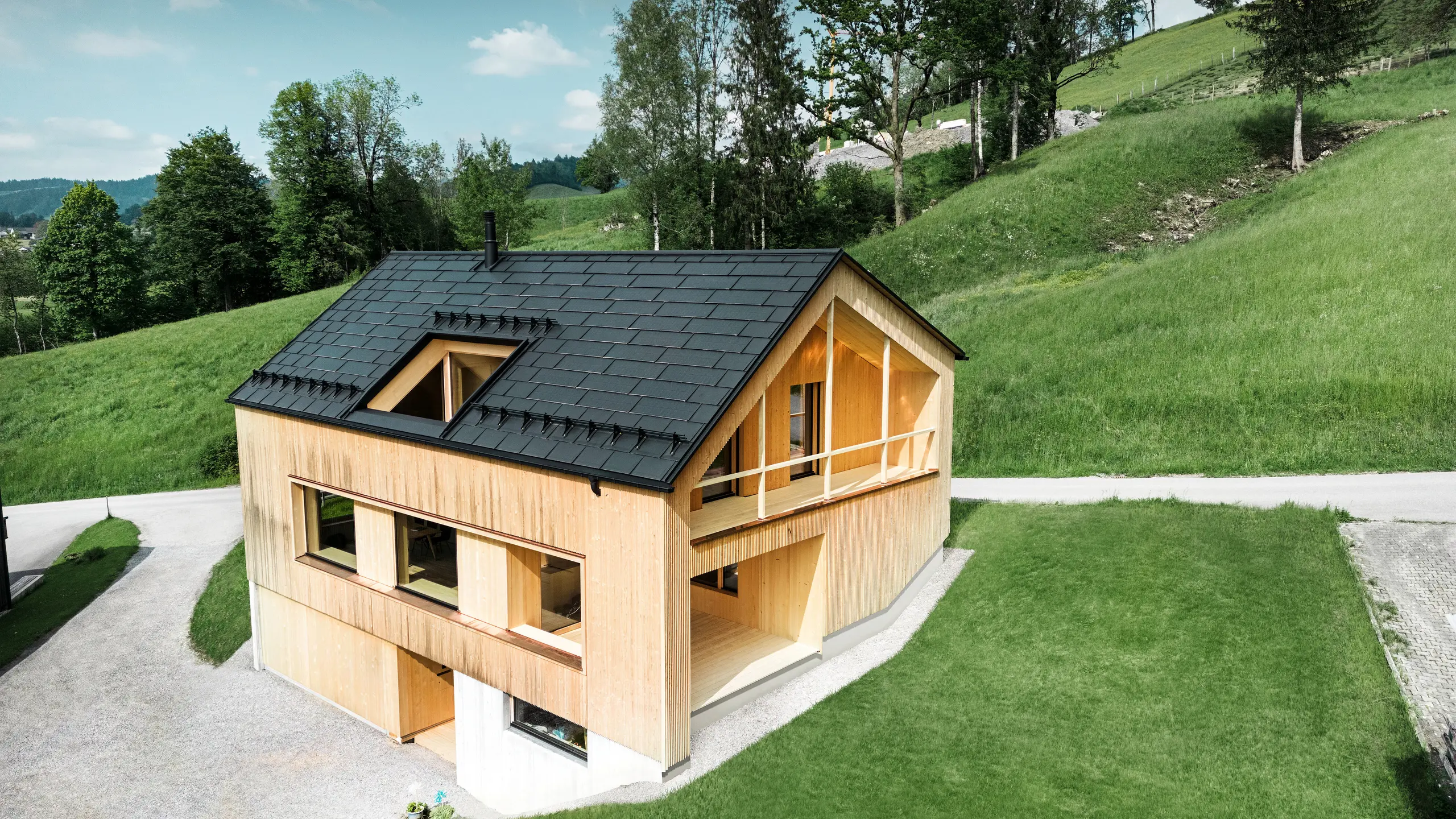 De foto toont een pittoresk ecohuis in Egg, genesteld tussen de glooiende groene heuvels, met een duurzaam PREFA-solar-dakpan. De zwarte, hoogwaardige PREFA-dakpan R.16 en de PREFA solar-dakpan passen harmonieus in het landelijke landschap en benadrukken de natuurlijke schoonheid van de omgeving. De solar-dakpan is niet alleen een blijk van toewijding aan hernieuwbare energie, maar ook een designelement dat de moderne houten gevel en de strakke lijnen van het gebouw stijlvol aanvult. Dit huis is een toonbeeld van milieubewust bouwen en een inspiratiebron voor toekomstgerichte architectuur.