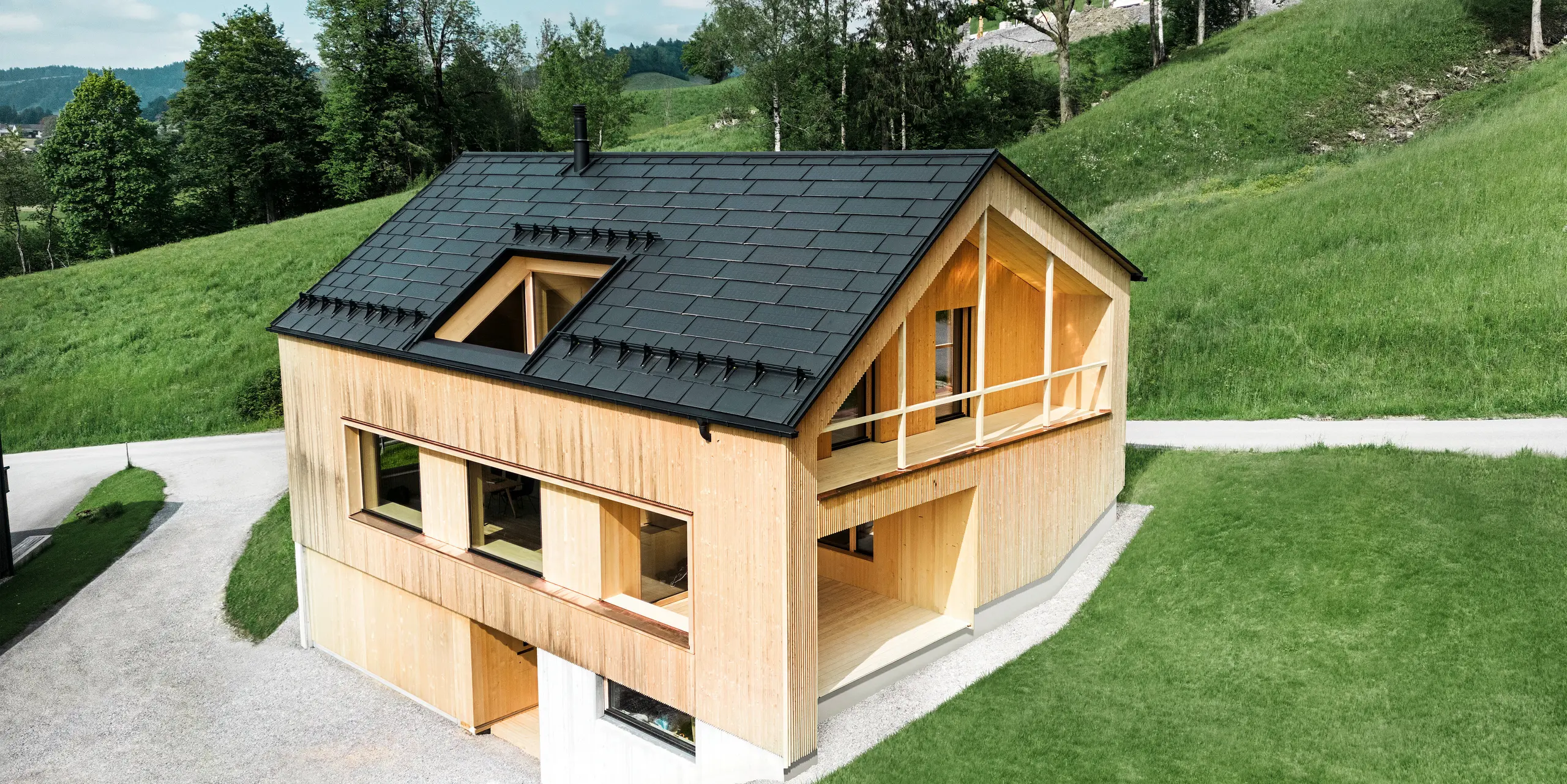 De foto toont een pittoresk ecohuis in Egg, genesteld tussen de glooiende groene heuvels, met een duurzaam PREFA-solar-dakpan. De zwarte, hoogwaardige PREFA-dakpan R.16 en de PREFA solar-dakpan passen harmonieus in het landelijke landschap en benadrukken de natuurlijke schoonheid van de omgeving. De solar-dakpan is niet alleen een blijk van toewijding aan hernieuwbare energie, maar ook een designelement dat de moderne houten gevel en de strakke lijnen van het gebouw stijlvol aanvult. Dit huis is een toonbeeld van milieubewust bouwen en een inspiratiebron voor toekomstgerichte architectuur.