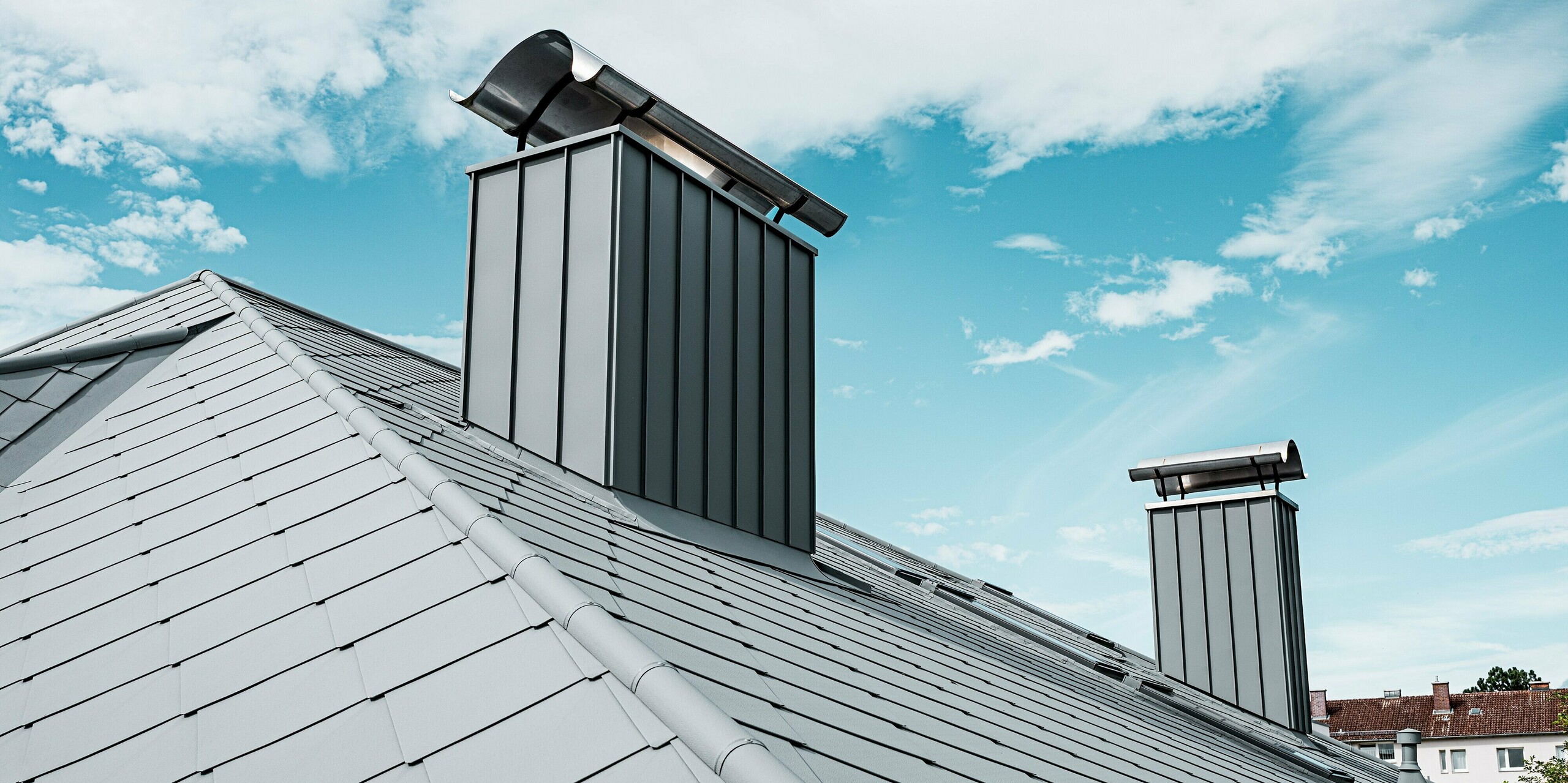 Das Bild zeigt die großflächige Dachfläche eines Einfamilienhauses, das mit PREFA Dachrauten 44 × 44 in P.10 Hellgrau eingedeckt ist. Die klaren Linien und die feine Stucco-Struktur verleihen dem Aluminiumdach eine moderne, elegante Optik. Zwei markant verkleidete Kaminzüge ragen aus der Dachfläche hervor und unterstreichen die hochwertige Ausführung der PREFA Aluminiumlösungen. Vor dem strahlenden Himmel erscheint das langlebige PREFA Dach besonders widerstandsfähig und präzise verarbeitet.