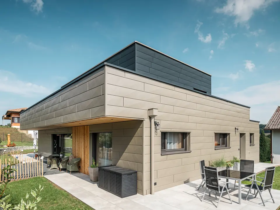 Une maison individuelle moderne à Wippenham, en Autriche, avec une façade en aluminium PREFA bicolore, composée des panneaux de façade Siding.X en bronze et anthracite. La façade présente des lignes et des bords élégants et clairement reconnaissables. Le système d'évacuation des eaux de toiture PREFA harmonieusement intégré avec collecteur d'eau et tuyau d'évacuation est clairement visible et s'intègre parfaitement dans le concept architectural global. Au premier plan, vous pouvez voir une agréable terrasse avec mobilier de jardin, qui souligne le design moderne de la maison nouvellement construite.