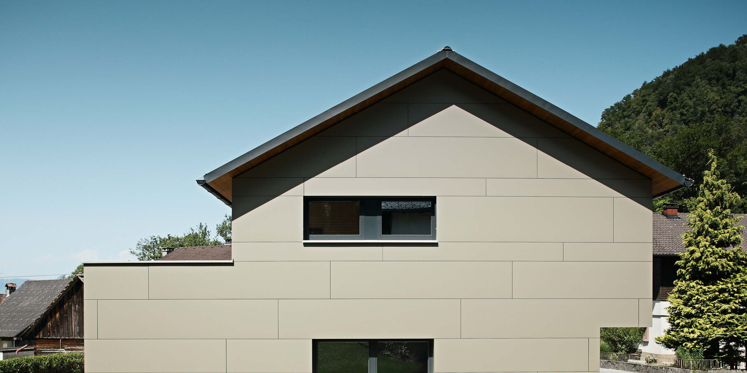 Frontansicht eines modernen Einfamilienhauses in Hohenems, das mit einem PREFA Aluminium Verbundplatten-Fassadensystem in Bronzemetallic verkleidet ist. Die Fassade zeigt eine harmonische Abfolge von horizontalen Linien, die eine ruhige und gleichzeitig zeitgenössische Ästhetik vermitteln. Das Haus fügt sich mit seiner zurückhaltenden Farbgebung und dem klaren Design nahtlos in die umgebende Landschaft ein, während das PREFA Dach – bestehend aus klassischen Dachplatten in P.10 Anthrazit – einen subtilen Kontrast bildet und den langlebigen Charakter des Gebäudes unterstreicht.