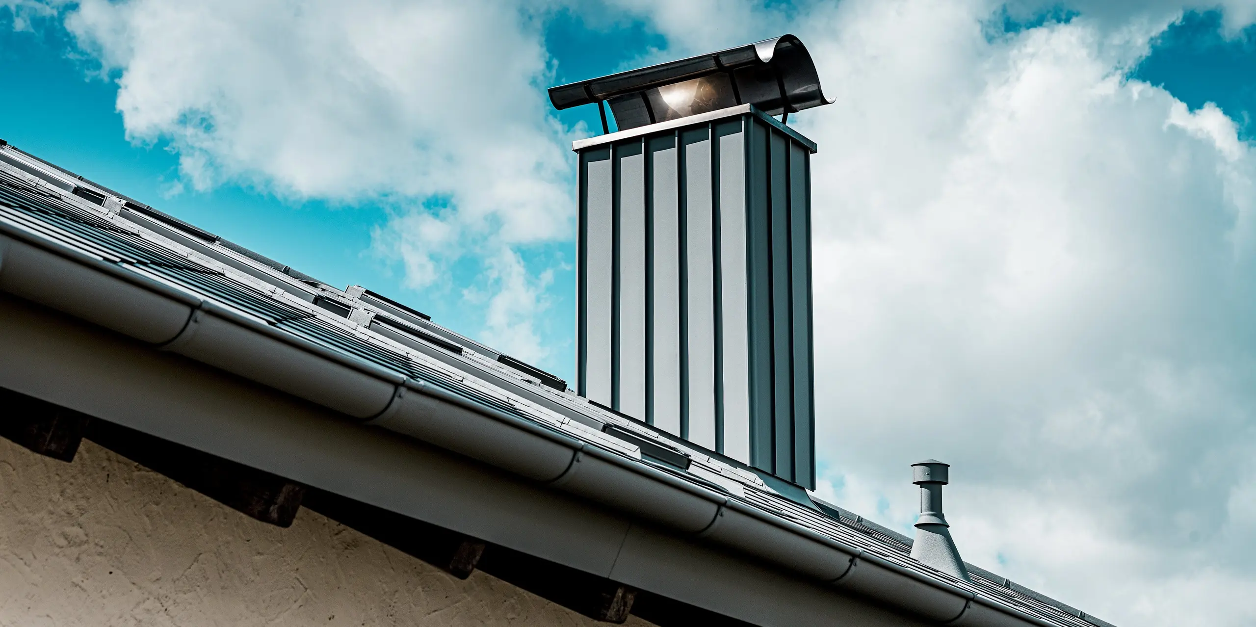 Das Bild zeigt eine Nahaufnahme des modern verkleideten Kamins eines Einfamilienhauses, der mit hochwertigem PREFA Aluminium in P.10 Hellgrau gestaltet ist. Die klaren vertikalen Linien der Kaminverkleidung harmonieren mit der robusten PREFA Dachfläche aus Dachrauten 44 × 44. Im Vordergrund ist die widerstandsfähige Aluminium-Dachrinne sichtbar, die sich nahtlos in das Dachsystem einfügt. Vor dem Hintergrund eines wolkigen Himmels hebt sich die langlebige, witterungsbeständige PREFA Lösung besonders markant ab.