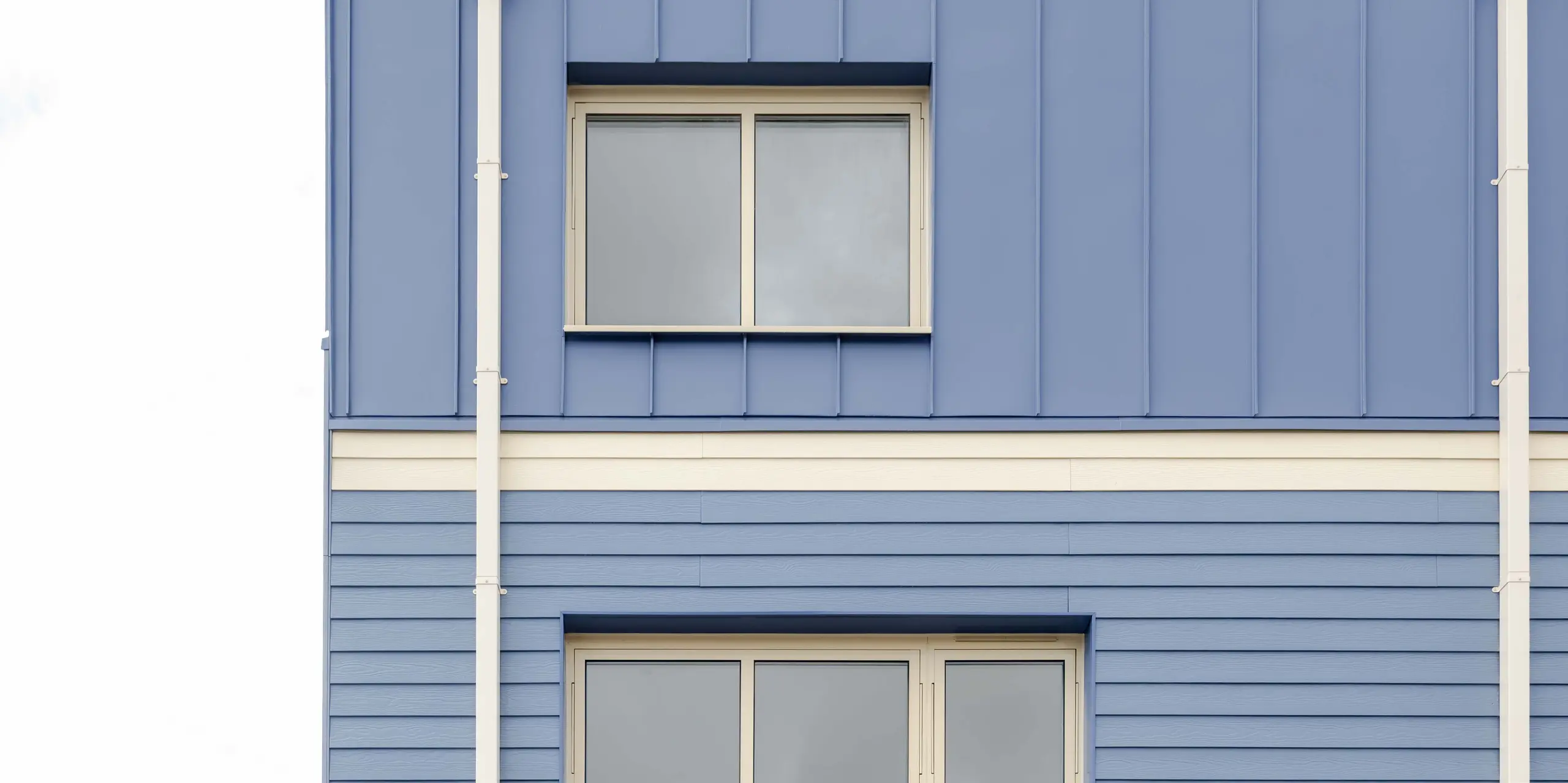 La photographie montre la façade arrière d'une maison mitoyenne de Goldsmith-Mews, revêtue du bardage PREFA FALZONAL bleu pigeon. Les plis verticaux en aluminium, à la structure bien définie, de la partie supérieure rejoignent les panneaux horizontaux de l'étage intermédiaire, créant une façade aux lignes affirmées. Les parties inférieures, de couleur claire et dotées de larges portes-fenêtres, ouvrent l'architecture sur le jardin. L'aluminium PREFA, de haute qualité, offre une excellente stabilité dimensionnelle et chromatique, une protection durable contre les intempéries et permet la réalisation d'une enveloppe de bâtiment moderne et précise, au caractère distinctif.