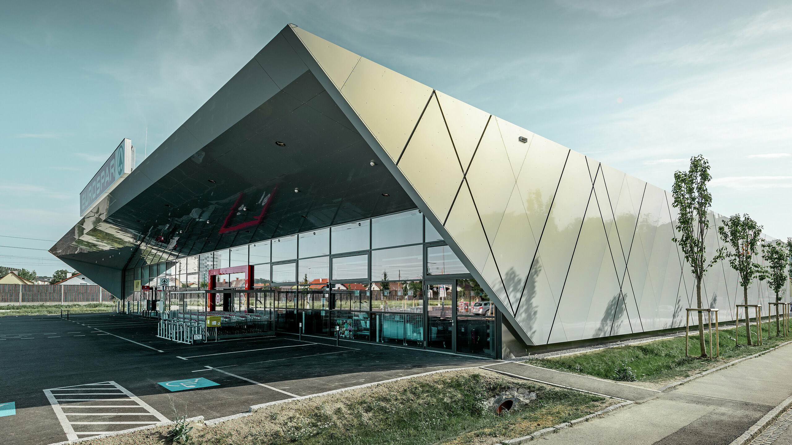 Entrée de la nouvelle filiale Eurospar de Wels habillée de panneaux de façade modernes de PREFA couleur argent doré ; selon l’angle de vue, à la lumière du soleil les panneaux composites en aluminium chatoient dans différentes couleurs.