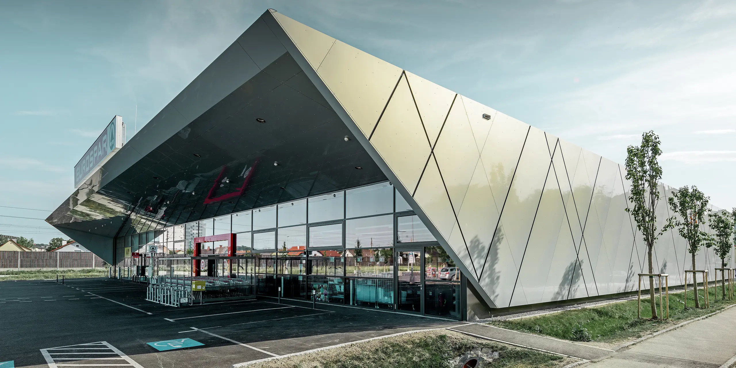 Der Eingangsbereich des EUROSPAR Supermarkts kombiniert großzügige Glasflächen mit einer markanten PREFA Aluminiumfassade. Die glänzende Oberfläche der Sonderfarbe Silver Gold unterstreicht die hochwertige, zukunftsorientierte Architektur der Supermarktfiliale. Die Aluminium Verbundplatten schillern in der Sonne je nach Blickwinkel in unterschiedlichen Farben.
