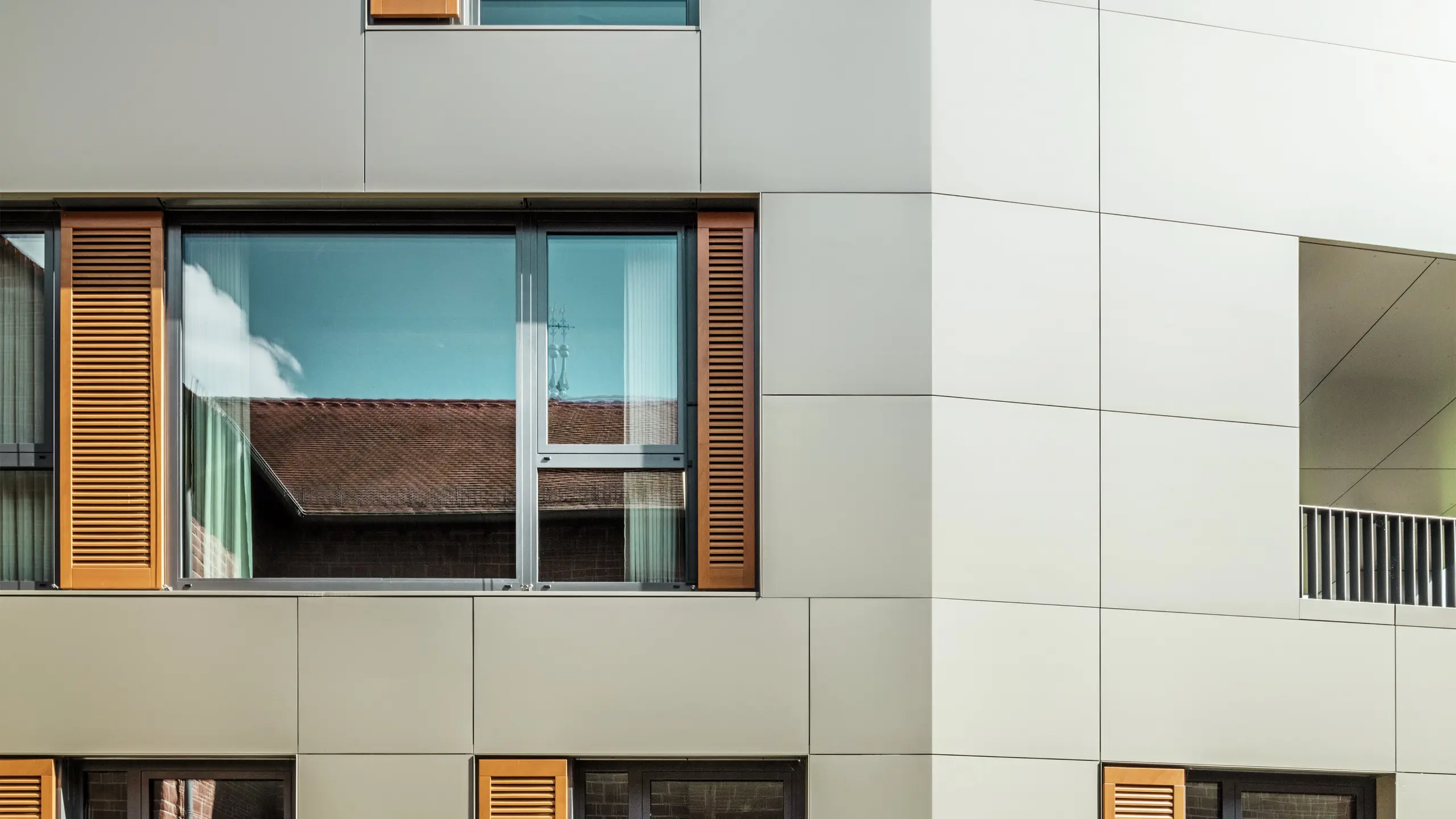 Detailaufnahme der Fassade des Rathauses in Alzenau, Deutschland, mit Aluminium-Verbundplatten von PREFA in der Farbe bronze. Die großen Fenster sind von eleganten Holzjalousien in warmem Braun umrahmt, die der Fassade eine ansprechende Textur verleihen. Im Vordergrund ist eine gepflegte Blumenrabatte zu sehen, die das urbane Design des Gebäudes mit grünen Akzenten ergänzt. Der klare Himmel und die saubere Linienführung der Architektur unterstreichen den modernen Charakter des Gebäudes.