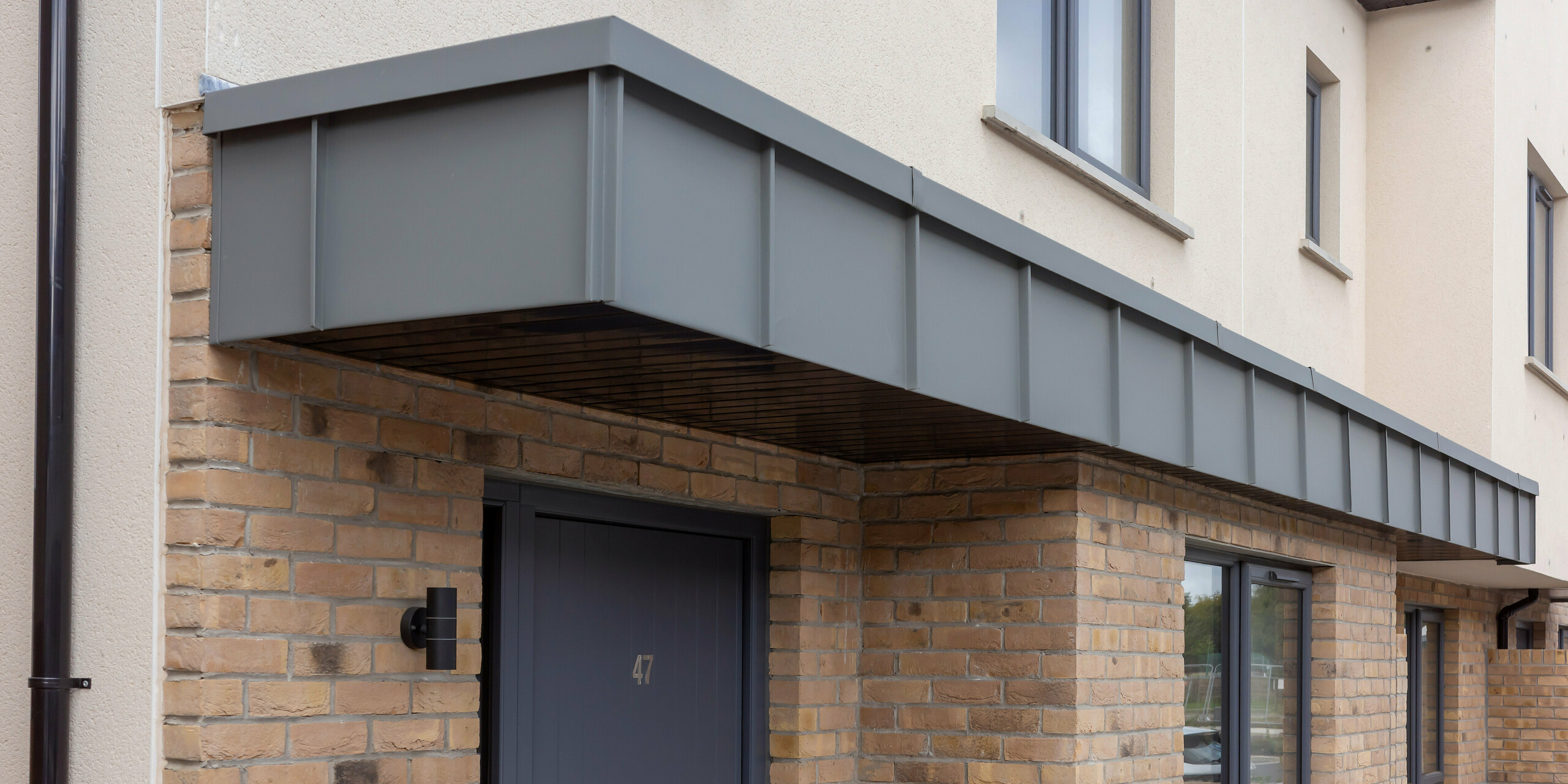 Die Detailaufnahme zeigt den Eingangsbereich eines Reihenhauses in Churchfields, bei dem das markante Vordach mit PREFALZ in P.10 Hellgrau verkleidet ist. Die klaren, geradlinigen Aluminiumflächen bilden einen modernen Kontrast zum warmen Backsteinmauerwerk und verleihen dem Eingang eine hochwertige, architektonische Tiefe. Die witterungsbeständige, farbstabile PREFA Oberfläche sorgt für dauerhaften Schutz und minimierten Wartungsaufwand – ideal für große Wohnanlagen. Das Zusammenspiel aus moderner Metalloptik und traditionellem Ziegelbau prägt die elegante Gesamtwirkung der Szene.