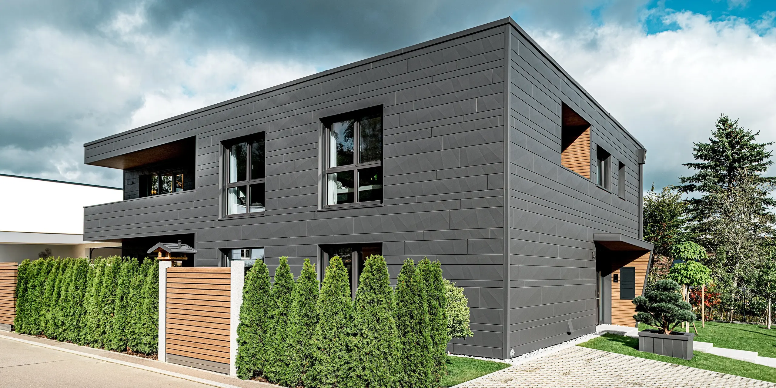 Vue latérale moderne d'une maison individuelle à Heidenheim, revêtue d'une façade en aluminium Siding.X PREFA couleur P.10 gris sombre. Les arêtes marquées et les largeurs variables des panneaux créent une texture dynamique. Devant la maison, une haie de thuyas dense guide le regard le long de la limite de propriété ; une clôture en bois offre un contraste chaleureux avec la froideur du métal. De larges baies vitrées et des niches habillées de bois renforcent le caractère moderne de la façade. Le revêtement PREFA, résistant aux intempéries, offre une excellente protection et un design de façade précis et intemporel.