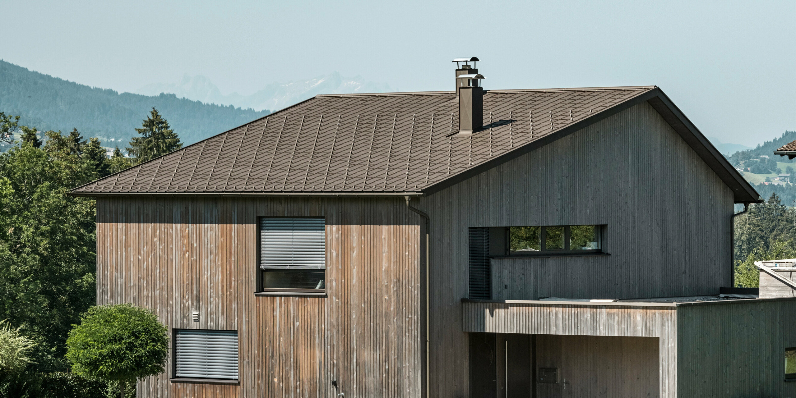 Vue latérale d'une maison individuelle contemporaine à Krumbach, avec une façade en bois naturel et une toiture en aluminium PREFA durable de couleur P.10 brun. L'harmonie des couleurs montre l'intégration élégante d'un design moderne et de matériaux traditionnels, tandis que les tuiles PREFA assurent une conception esthétique et fonctionnelle de la toiture.