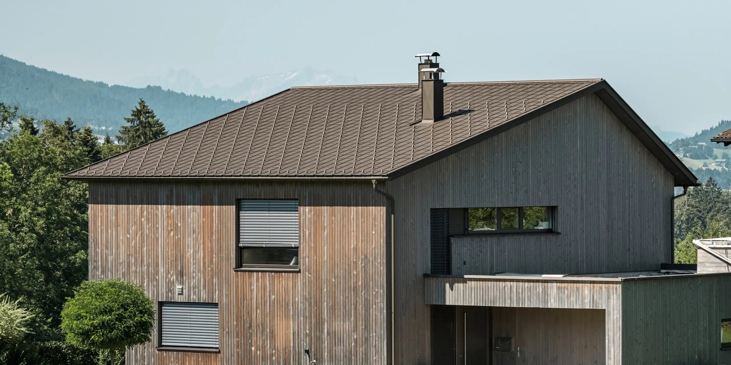 Zijaanzicht van een moderne vrijstaande woning in Krumbach met een combinatie van een gevel van natuurlijk hout en een duurzaam PREFA aluminium dak in P.10 bruin. De harmonieuze kleurstelling toont de elegante integratie van modern design en traditionele materialen, waarbij de PREFA- dakpannen voor een esthetisch en functioneel dakdesign zorgen.