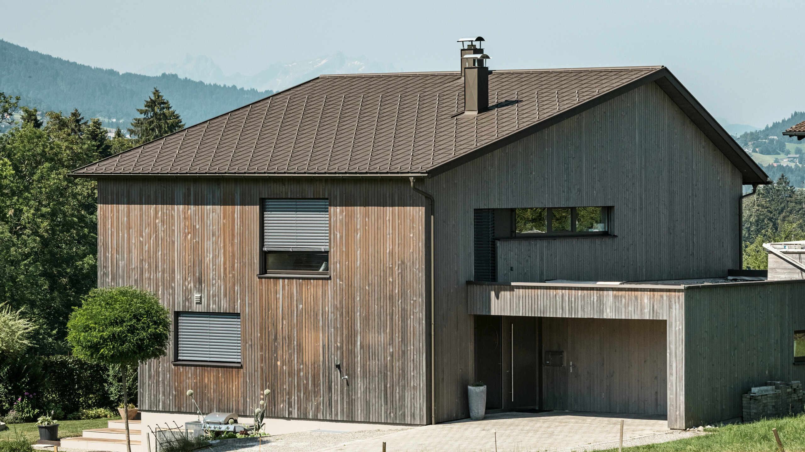 Vue latérale d'une maison individuelle contemporaine à Krumbach, avec une façade en bois naturel et une toiture en aluminium PREFA durable de couleur P.10 brun. L'harmonie des couleurs montre l'intégration élégante d'un design moderne et de matériaux traditionnels, tandis que les tuiles PREFA assurent une conception esthétique et fonctionnelle de la toiture.
