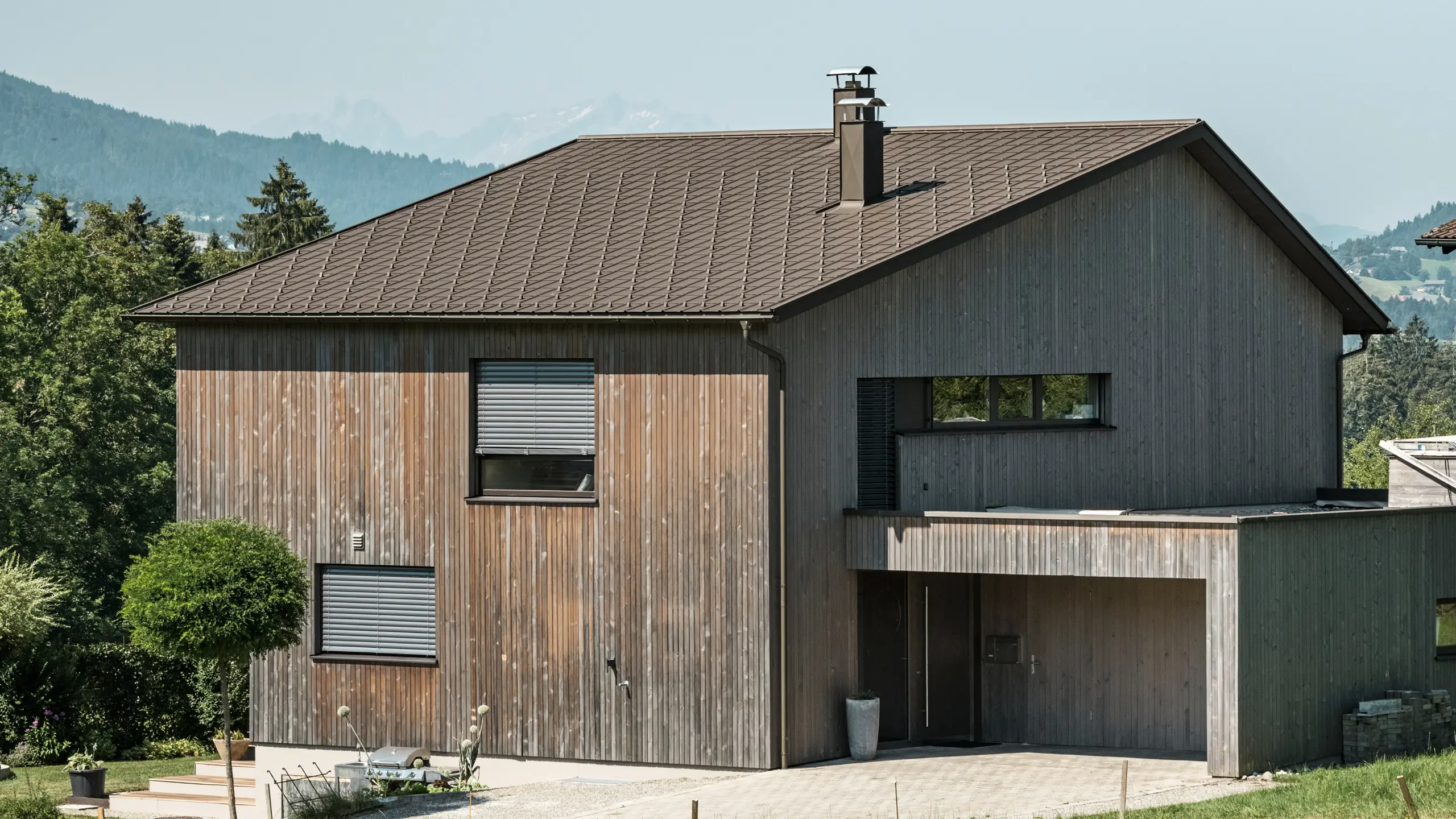 Vue latérale d'une maison individuelle contemporaine à Krumbach, avec une façade en bois naturel et une toiture en aluminium PREFA durable de couleur P.10 brun. L'harmonie des couleurs montre l'intégration élégante d'un design moderne et de matériaux traditionnels, tandis que les tuiles PREFA assurent une conception esthétique et fonctionnelle de la toiture.