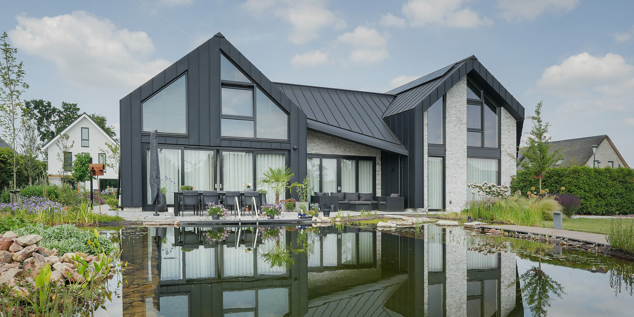 Idyllisches Bild eines Familienhauses in Veenendaal, Niederlande, das sich in einem klaren Gartenteich spiegelt. Das dunkle PREFALZ Dach- und Fassadensystem kontrastiert mit dem hellblauen Himmel, während der Teich das Haus und die umgebende Natur spiegelt und die friedliche Atmosphäre unterstreicht. Ein gepflegter Garten mit einer Vielzahl von Pflanzen und Sitzgelegenheiten lädt zum Verweilen ein und vervollständigt das Bild einer harmonischen Wohnästhetik in Verbindung mit der Natur.