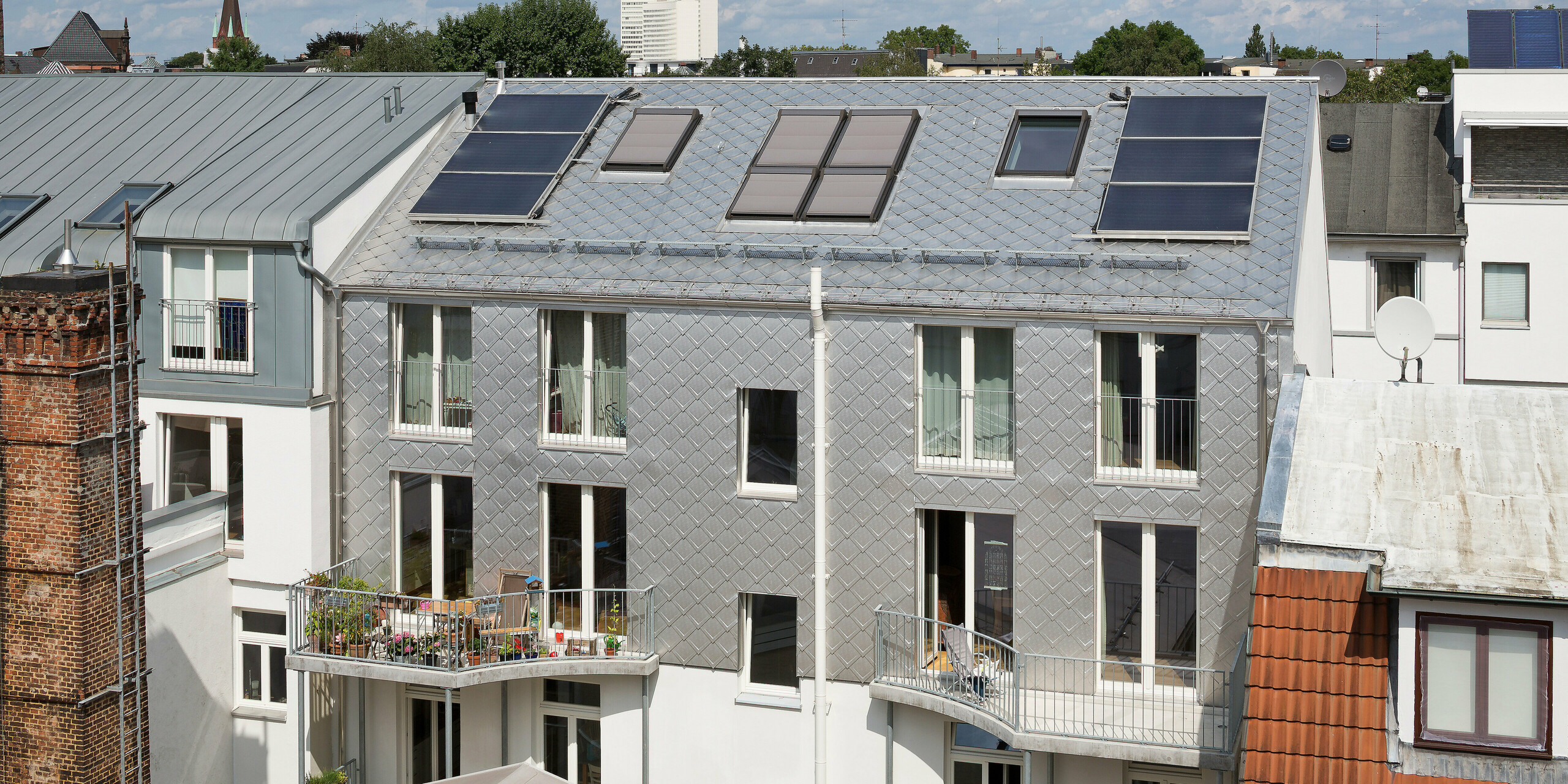 Das Dach eines eleganten Wohngebäudes in Hamburg besticht durch die Verwendung der PREFA Dachraute 29 × 29 in Naturblank. Die Rautenstruktur hebt sich beeindruckend von der Umgebung ab. Solarzellen auf dem Dach ergänzen das nachhaltige Design, während die Balkone der unteren Etagen das Gebäude noch einladender wirken lassen.