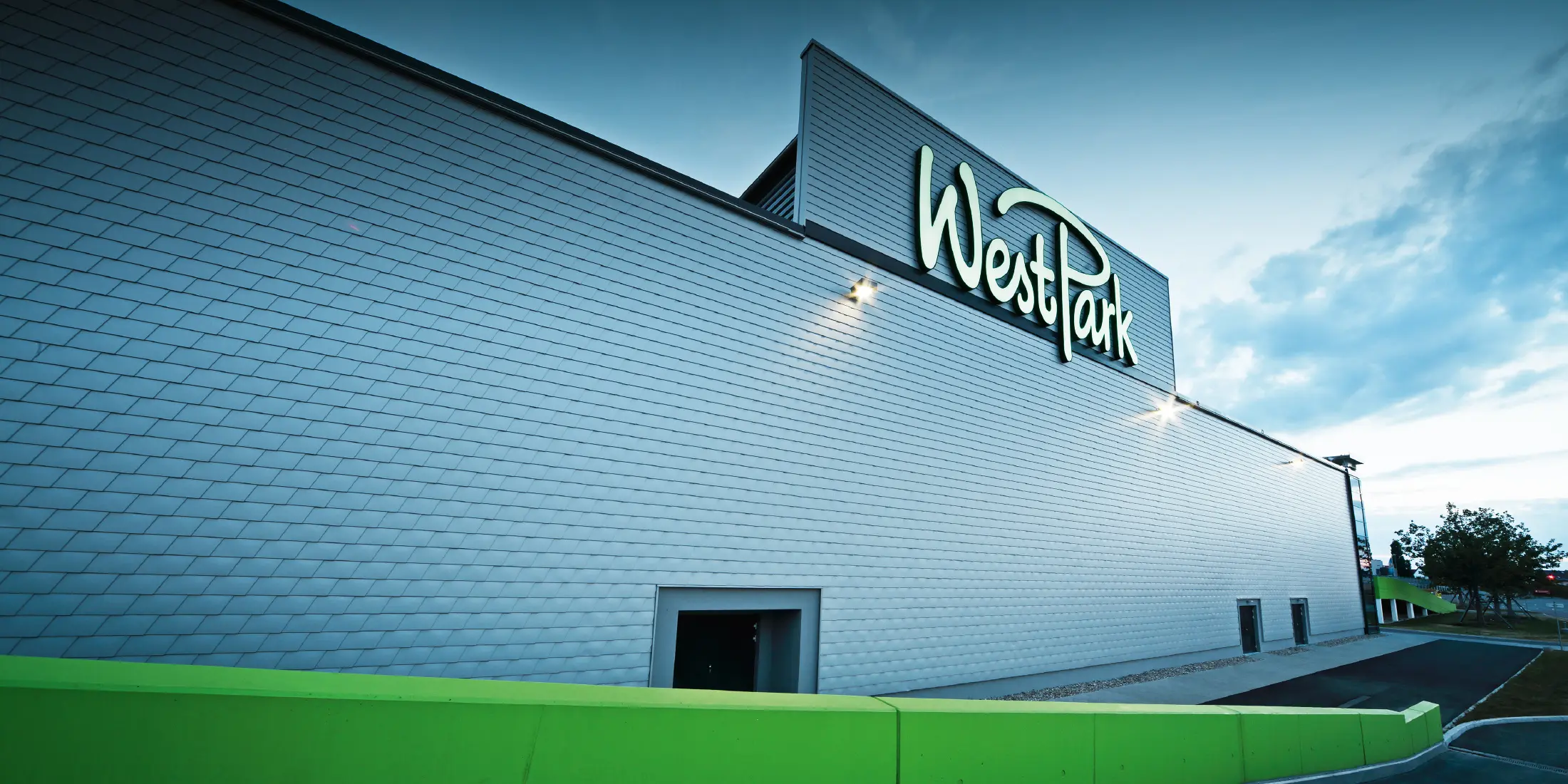 Moderne Fassade eines Einkaufszentrums mit großem Schriftzug „WestPark“. Die Außenwand ist mit silbermetallicfarbenen Aluminium-Schindeln verkleidet und wirkt durch die gleichmäßige Struktur elegant und technisch. Im Vordergrund verläuft eine leuchtend grüne Mauer entlang des Gehwegs. Das Gebäude ist in der Abenddämmerung beleuchtet.