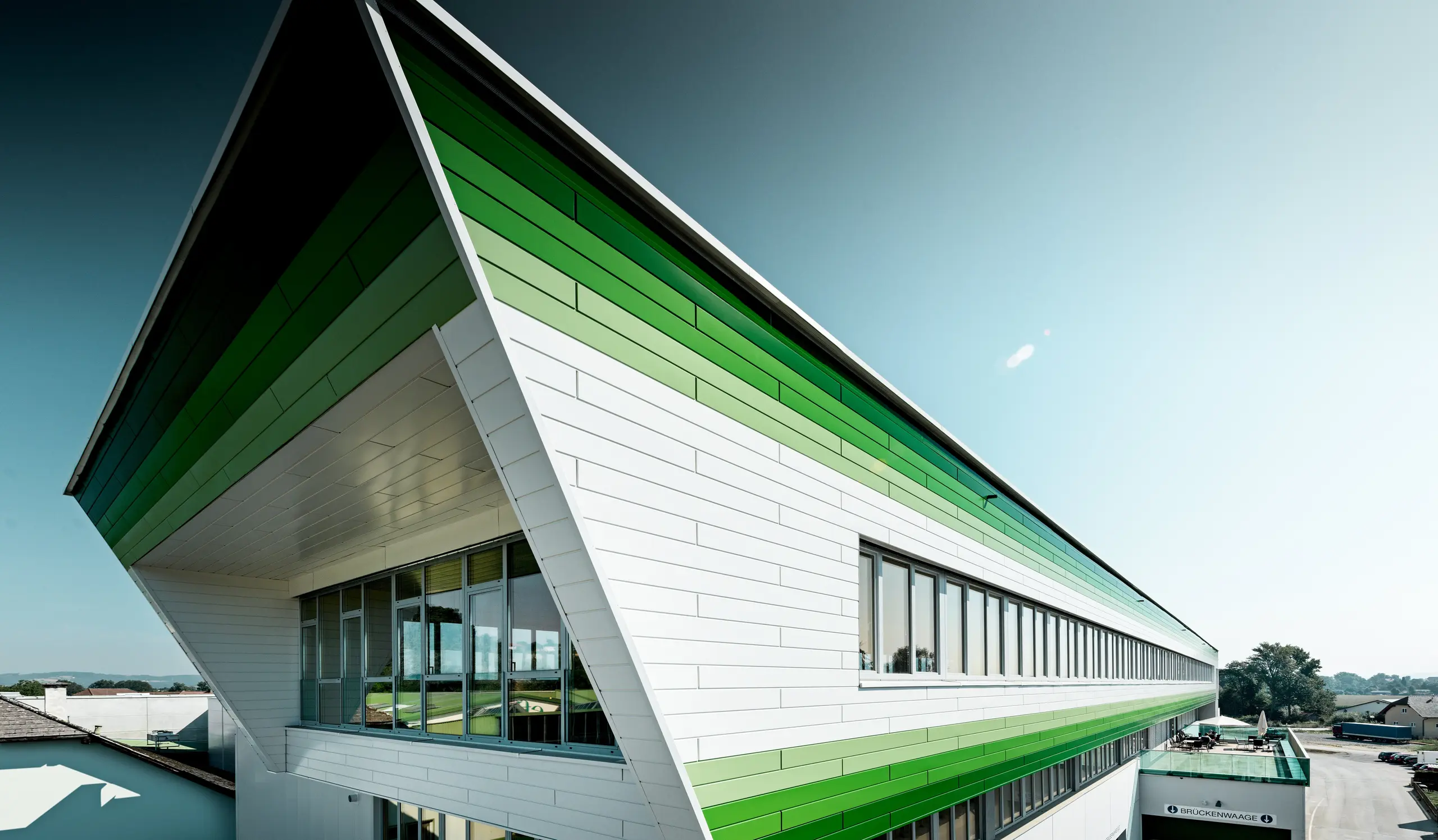 Weitläufige Ansicht des Firmengebäudes der efko Frischfrucht und Delikatessen GmbH mit markanter PREFA Siding-Fassade in P.10 Prefaweiß und individuellen grünen RAL-Sonderbeschichtungen. Die horizontalen Aluminiumprofile betonen die außergewöhnliche Gebäudelänge und schaffen ein dynamisches Fassadenbild. PREFA Aluminium steht für langlebige Qualität, hohe Witterungsbeständigkeit, Farbstabilität und eine wartungsarme Lösung für moderne Unternehmensarchitektur.