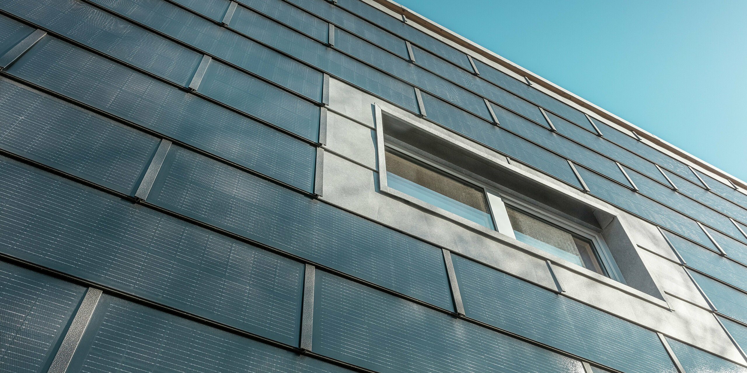 Vue oblique de la façade de l'entreprise de couverture RÜMA, dans le Vorarlberg, revêtue de tuiles solaires PREFA. Les éléments en aluminium gris et les modules photovoltaïques intégrés encadrent une fenêtre. Un ciel bleu éclatant apparaît en arrière-plan.