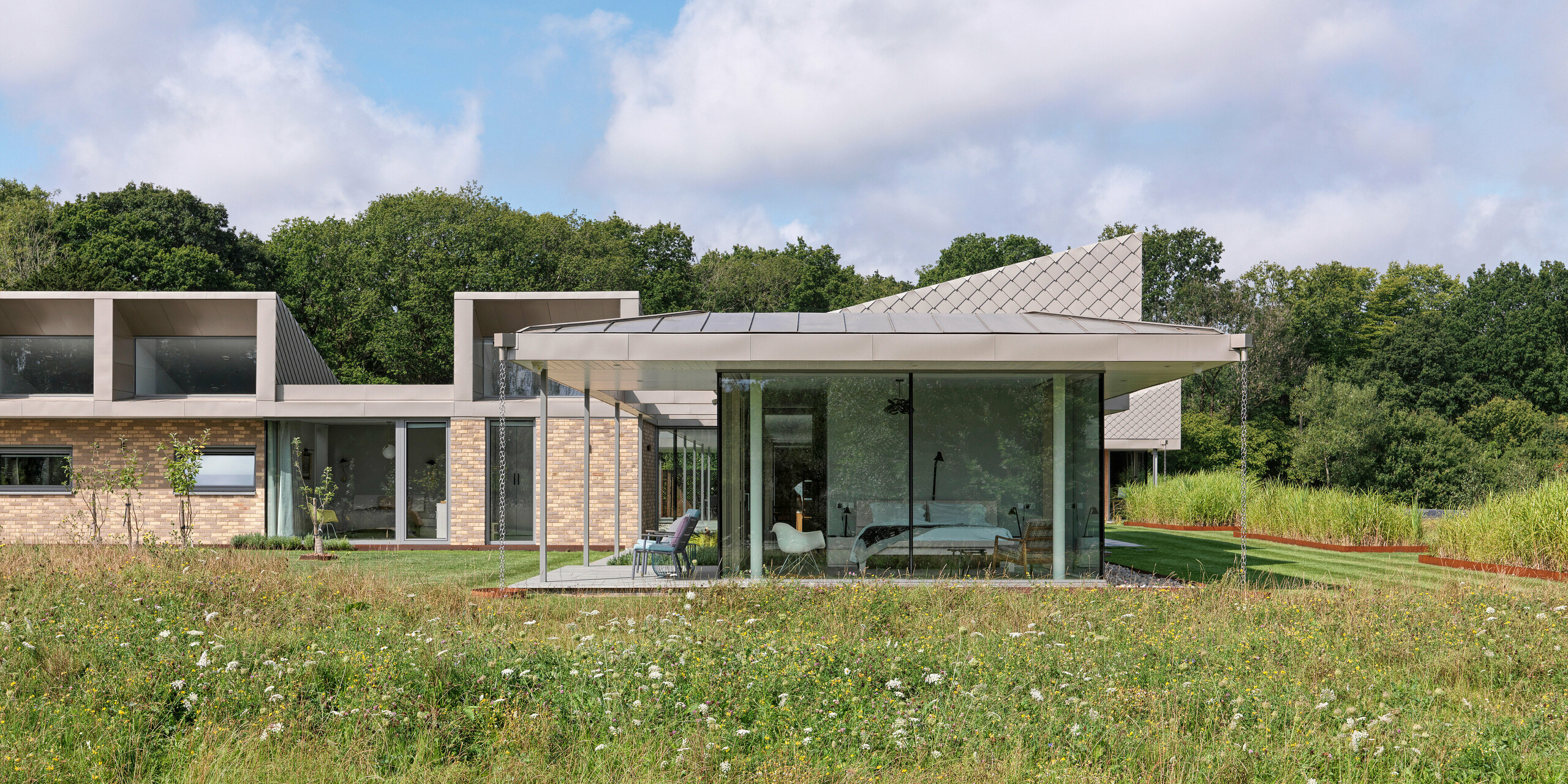 Vue latérale d'un bungalow moderne dans le West Sussex, doté d'une toiture en aluminium PREFALZ et de façades accentuées en losanges muraux PREFA 20 × 20 en P.10 bronze. Les baies vitrées des pièces à vivre s'ouvrent sur la prairie fleurie au premier plan, créant une connexion harmonieuse entre l'intérieur et la nature. Le système de toiture et de façade PREFA, résistant, assure non seulement la protection du bâtiment, mais lui confère également une élégance architecturale.