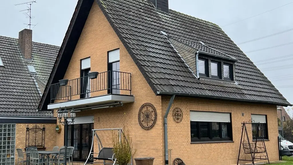 Maison individuelle en briques jaunes avec toit en pente raide et couverture de toit sombre avant la rénovation du toit ; côté jardin, avec balcon, lucarne, terrasse avec mobilier et pelouse au premier plan.