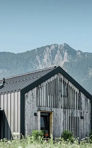 Moderner, funktionaler Wohnbau mit einer Kombination aus Holzfassade und PREFA Band- und Blechmaterial für das Dach. Die klare Architektur wird durch die elegante, metallische Dach- und Fassadenverkleidung unterstrichen, die ideal für anspruchsvolle Projekte ist. Das Bild veranschaulicht die vielseitigen Anwendungsmöglichkeiten der PREFA Produkte im Bereich Bänder und Bleche.