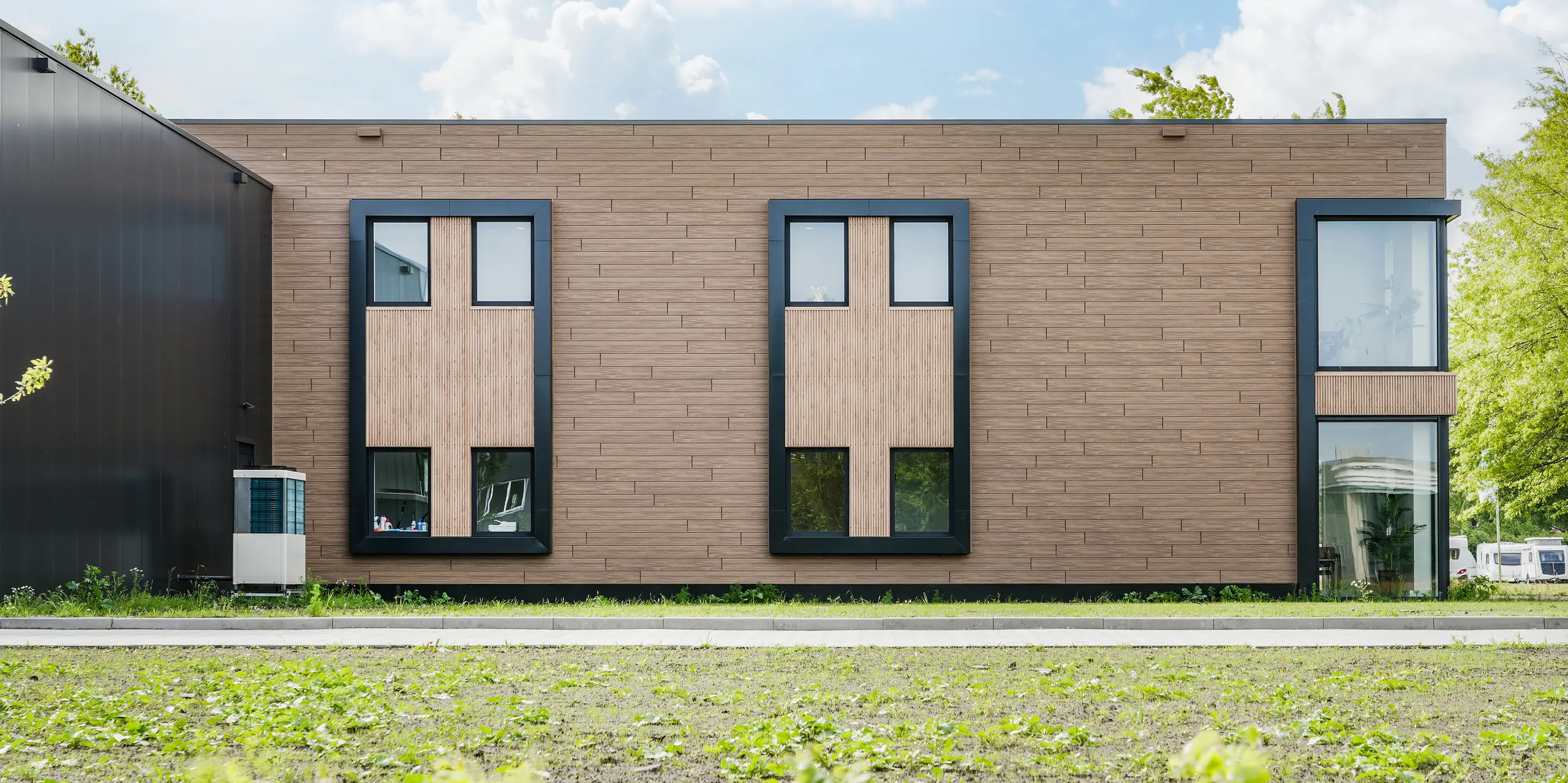 Die geradlinige Gebäudefassade der Dokter Schoonmaakorganisatie in Wolvega beeindruckt mit einer ruhigen, modernen Gestaltung und markanten Fensterelementen. Die walnussbraune PREFA Fassade mit Sidings in Holzoptik verleiht dem Baukörper eine warme, natürliche Ausstrahlung. Dunkle Rahmen setzen spannende Kontraste und betonen die hochwertige Architektur des Unternehmensgebäudes.