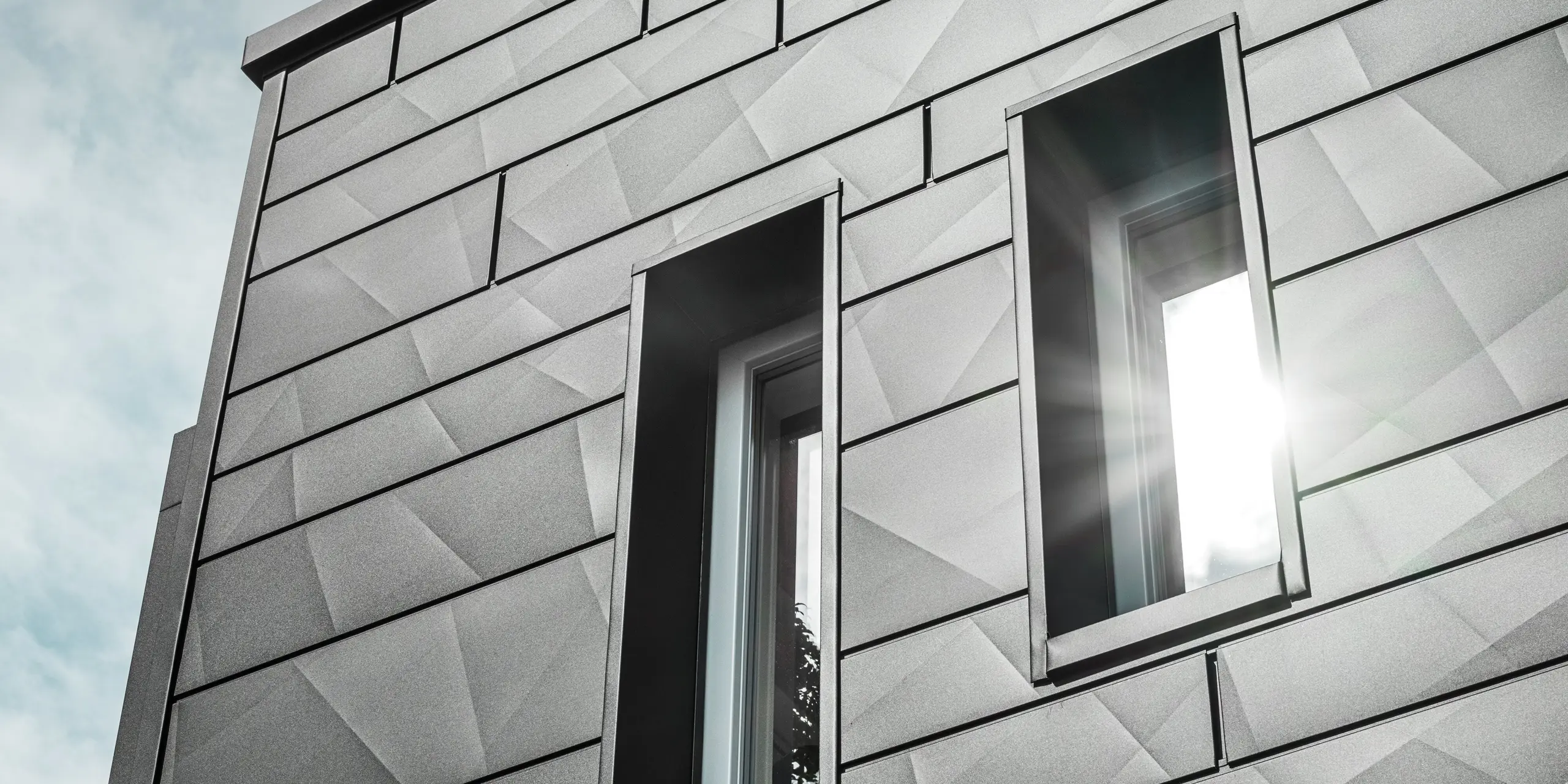 Die moderne Fassade des sanierten Einfamilienhauses in Teuschnitz zeigt die geometrischen PREFA Siding.X Fassadenpaneele in P.10 Schwarz. Die Fensterumrahmungen aus schwarzen Aluminium Verbundplatten fügen sich harmonisch in das Gesamtbild ein. Ein Sonnenstrahl fällt durch eines der Fenster und betont die klare, strukturierte Oberfläche der Fassadenpaneele. Die ästhetische Wirkung der Fassade wird durch das subtile Zusammenspiel von Licht und Material verstärkt.