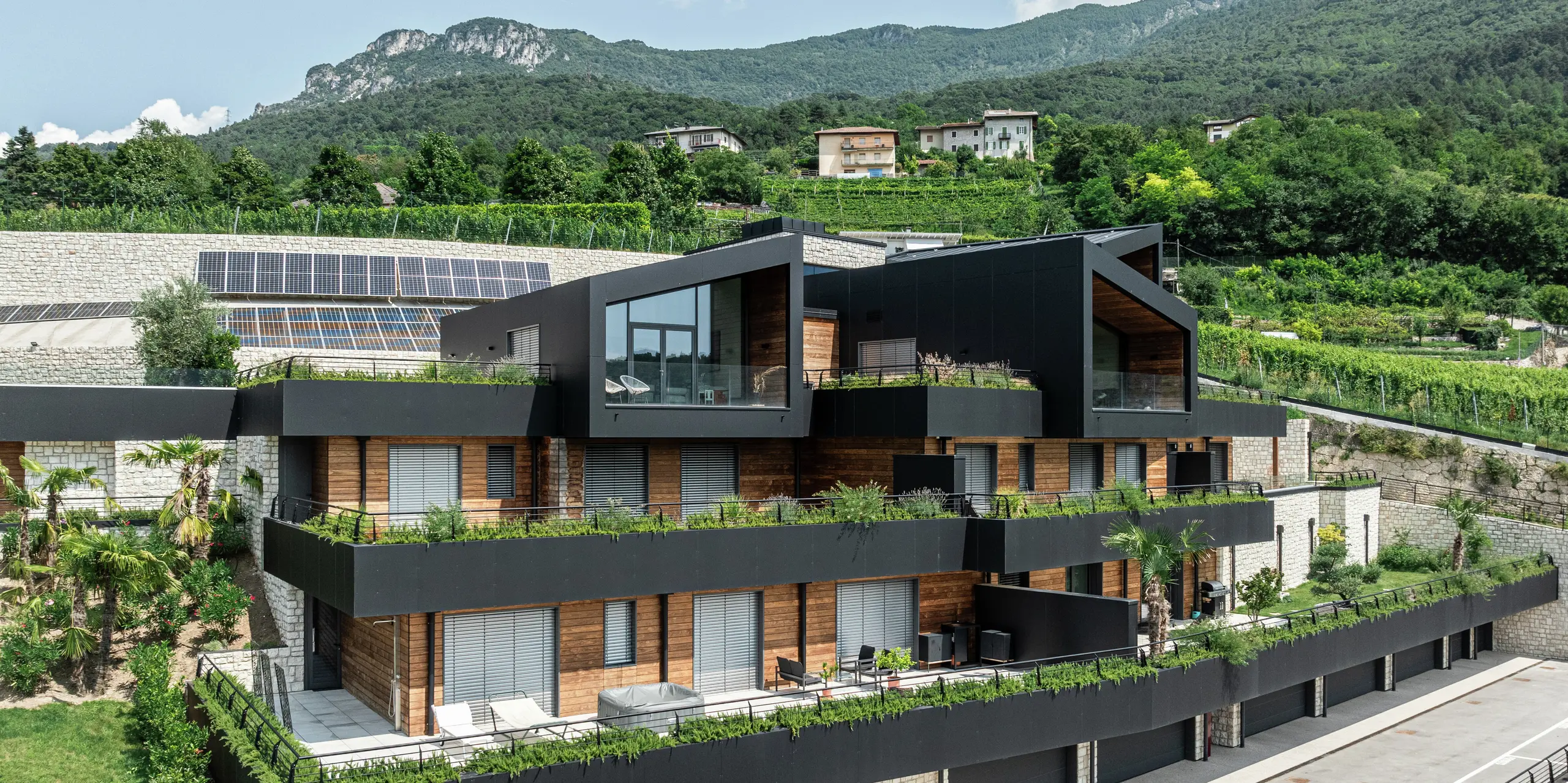 [Translate to Français:] Moderne Wohnanlage in Hanglage mit nachhaltigem Design: Die Kombination aus Naturstein, Holz und der hochwertigen PREFA Aluminiumfassade in P.10 Schwarz verleiht dem Gebäude eine elegante und zeitlose Optik. Das PREFALZ Dach in P.10 Schwarz bietet optimalen Schutz und Langlebigkeit. Großzügige Terrassen, begrünte Dächer und Photovoltaikanlagen unterstreichen das nachhaltige Wohnkonzept. Die harmonische Einbettung in die Landschaft sowie die hochwertigen und pflegeleichten PREFA Materialien garantieren eine stilvolle und witterungsbeständige Architektur mit höchstem Wohnkomfort.