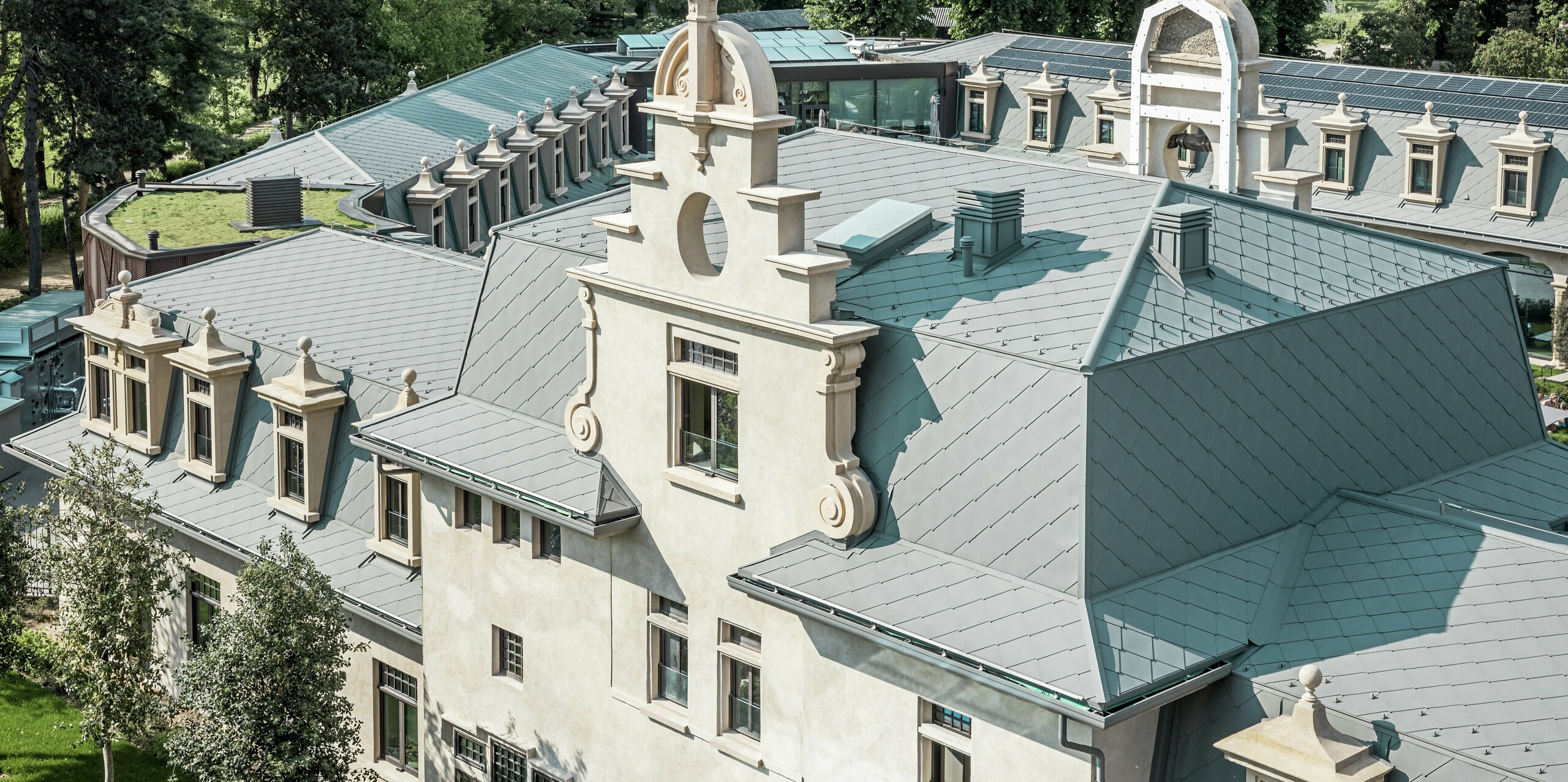 Vue détaillée du toit du parc thermal « De Montel – Terme Milano », avec sa toiture finement réalisée en losanges PREFA 44 × 44 P.10 gris souris. La géométrie complexe du toit, avec ses nombreuses lucarnes, corniches, pignons et éléments de ventilation, est magistralement exécutée. L'architecture historique est harmonieusement complétée par des éléments modernes, tels qu'une toiture plate végétalisée et le système photovoltaïque parfaitement intégré en arrière-plan.