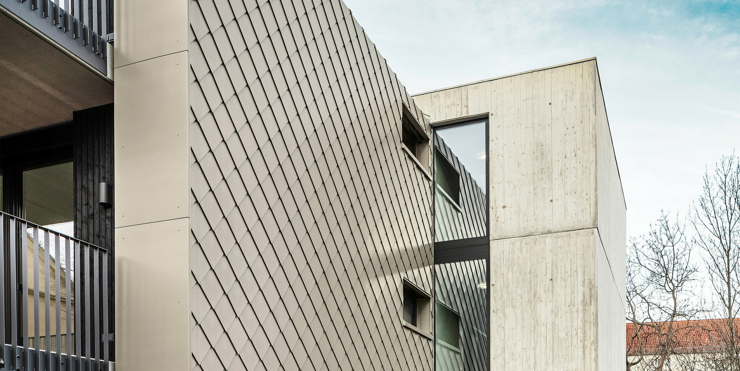 Zugangsbereich zu einem Mehrfamilienhaus in der Chemnitzer Straße im Bezirk Plauen, Dresden. Das Gebäude wird geschützt von einem robusten PREFA Dach- und Fassadensystem aus Aluminium. Die hochwertigen PREFA Dach- und Wandrauten 29 × 29 in Bronze sorgen für eine elegante Optik und langlebigen Schutz. Das Bild zeigt den Eingangsbereich, der von der eleganten Rautenstruktur und einer Betonwand flankiert wird. Die architektonische Gestaltung kombiniert robuste Materialien mit klaren Linien und modernen Elementen, was dem Gebäude eine zeitgemäße und stilvolle Optik verleiht.
