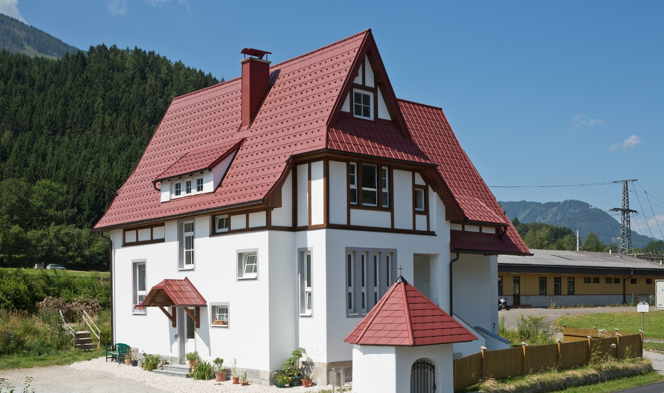 Verwinkeltes Steildach mit PREFA Dachplatte in P.10 Oxydrot mit vielen Details wie Gauben, Türmchen und Kamineinfassungen. Auch das Vordach und das Marterl vor dem Haus sind mit PREFA Blechdach eingedeckt.