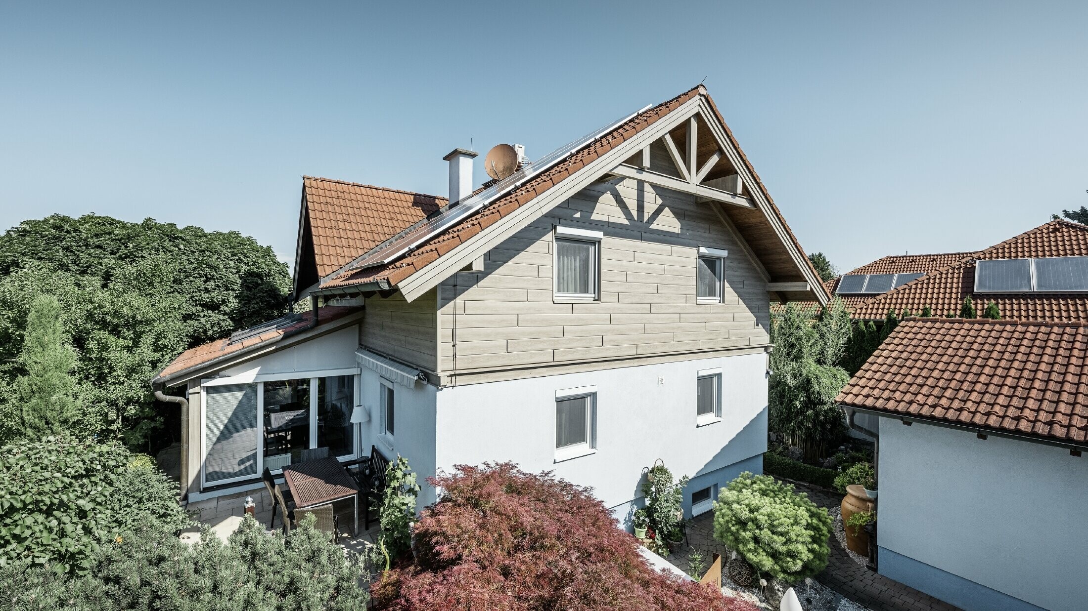 Einfamilienhaus mit Satteldach und roter Dacheindeckung, kombiniert mit einer modernen Fassadengestaltung. Der obere Giebel ist mit beige-grauen Aluminium-Fassadenpaneelen von PREFA verkleidet, während das Erdgeschoss eine weiße Putzfassade hat. Auf dem Dach befinden sich Solarmodule. Das Haus liegt in einem grünen Garten mit Terrasse, Gartenmöbeln und üppiger Bepflanzung, umgeben von weiteren Wohnhäusern mit ähnlicher Dacharchitektur.