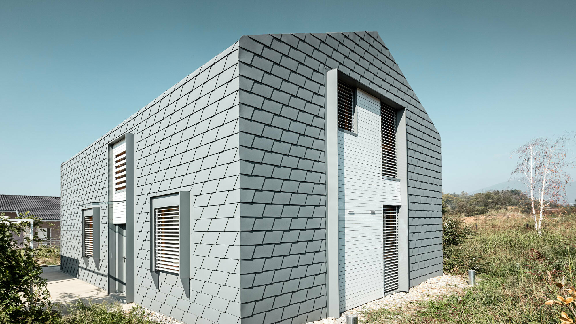 Vue de trois quarts de la maison avec espaces verts. La couverture ininterrompue en bardeaux et les stores extérieurs sont visibles.