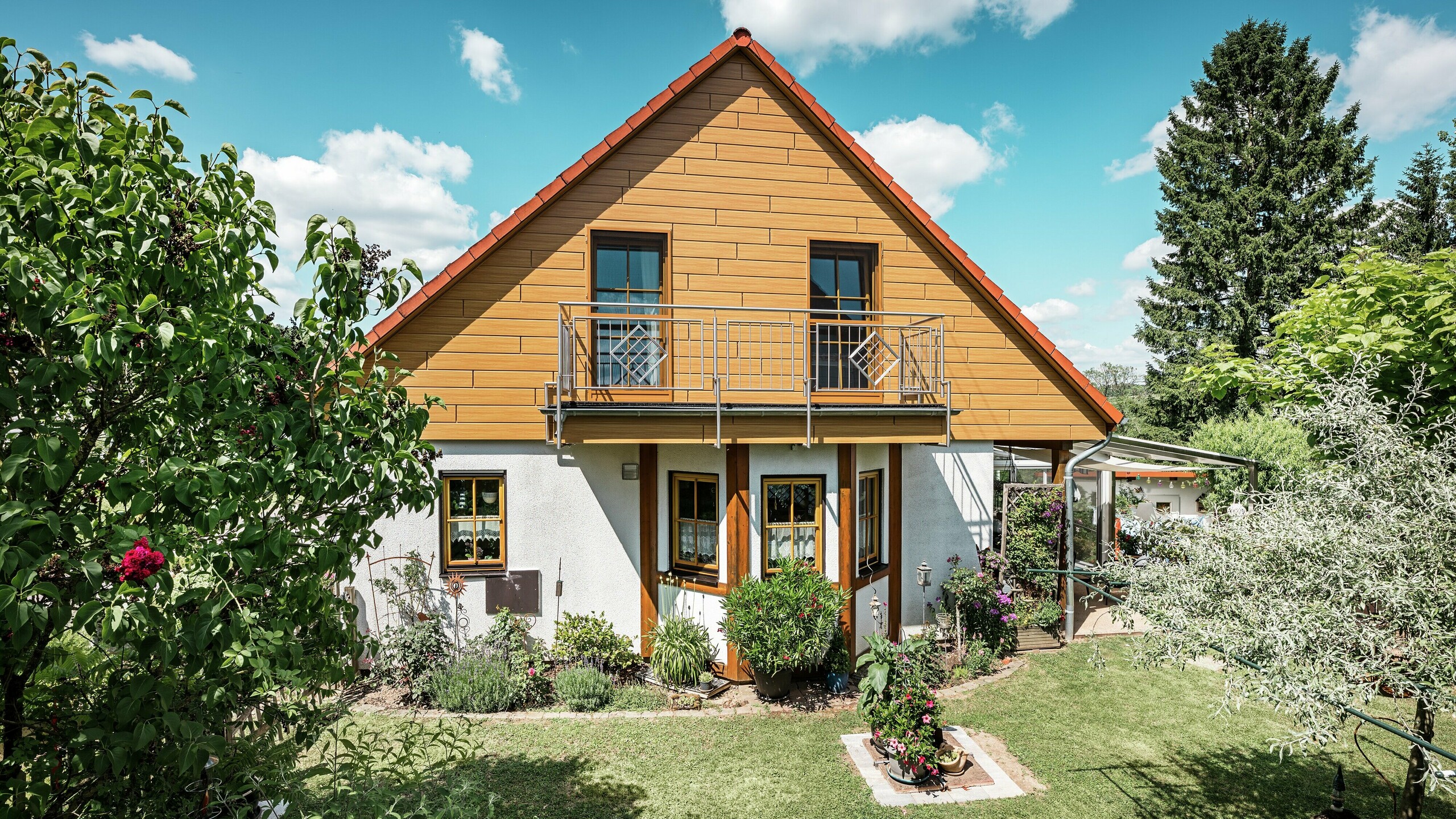 Op deze foto straalt de gerenoveerde gevel van een eengezinswoning in Blieskastel in een warme, natuurlijke eiken naturel-kleur dankzij de PREFA Siding. De hoogwaardige aluminium gevel met realistische houtlook zorgt voor een sterk visueel accent en sluit indrukwekkend aan bij het rode pannendak en de weelderige tuin. Omgeven door bloemen, struiken en weelderig groen oogt het huis uitnodigend en verzorgd. Het PREFA-oppervlak combineert een natuurlijke uitstraling met functionele voordelen zoals hoge weersbestendigheid, stabiliteit en een vrijwel onderhoudsvrije levensduur – een esthetisch hoogtepunt met duurzame meerwaarde.