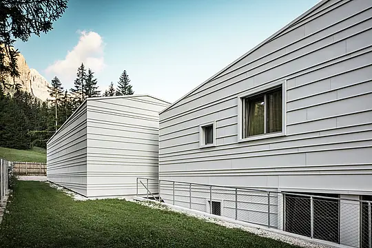 Die Rückseite der „Tempesti“-Kaserne in Corvara bietet einen beeindruckenden Ausblick: Wald, strahlend blauer Himmel und sonnenbeschienene Berggipfel prägen das Bild. Die helle Aluminiumfassade mit ihrer klaren Struktur spiegelt die technische Raffinesse von PREFALZ in der Farbe P.10 Prefaweiß wider. Die horizontalen Aluminiumstreifen reflektieren das Licht, bieten optimalen Witterungsschutz und fügen sich harmonisch in die Alpenlandschaft ein. Ein Paradebeispiel für langlebige und moderne Militärarchitektur!