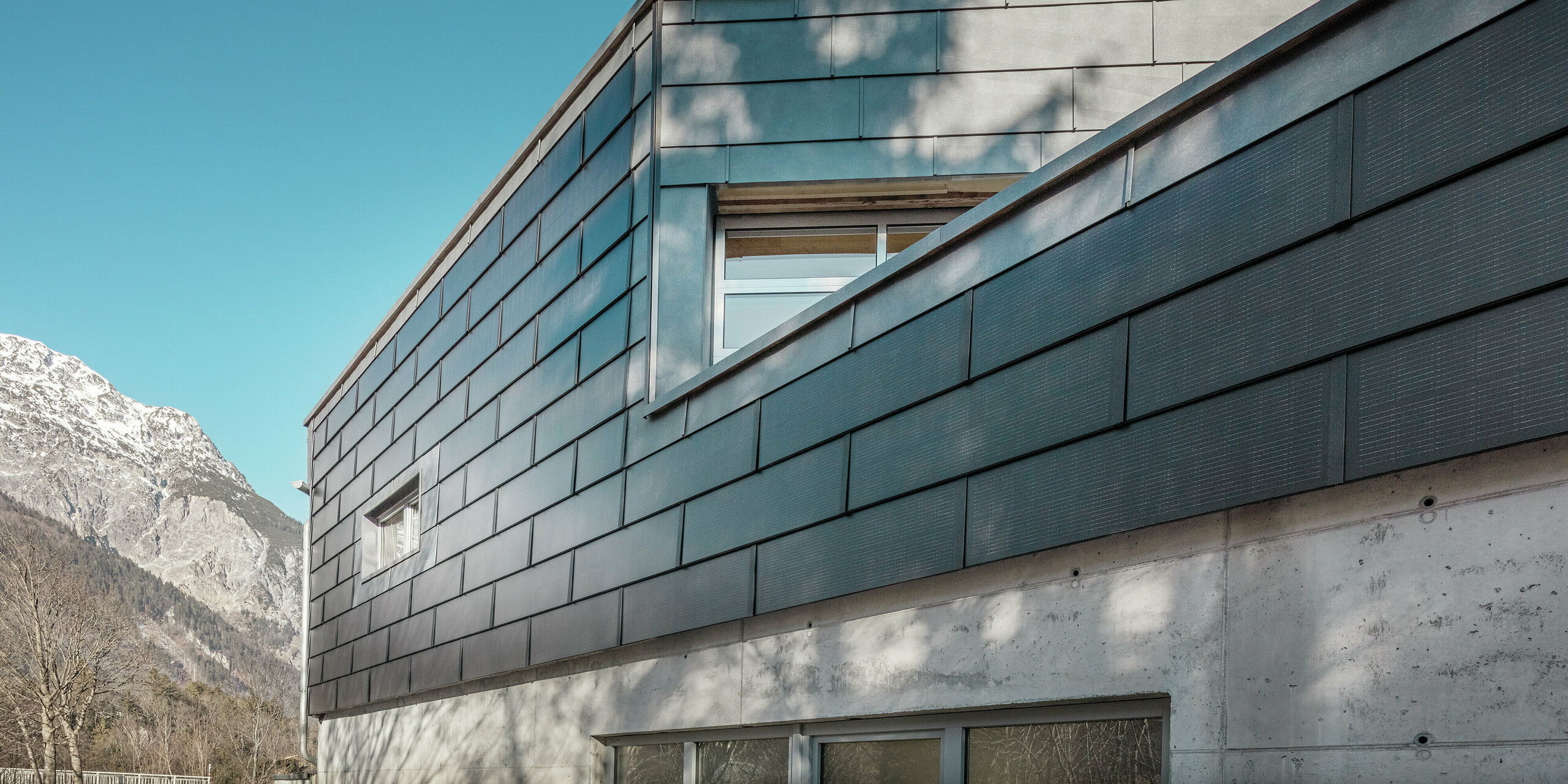 Façade du bâtiment de l'entreprise de métallurgie RÜMA dans le Vorarlberg avec des tuiles solaires PREFA innovantes et du béton apparent, entourée de montagnes sous un ciel bleu clair, avec des ombres d'arbres sur le mur.