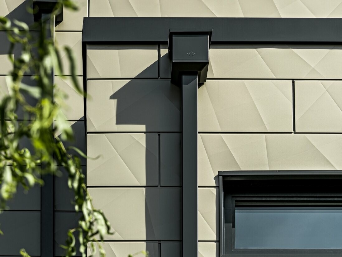 Maison individuelle moderne avec toit plat et fenêtres fines. La façade est revêtue du Siding.X de façade PREFA style plissé, dans la couleur bronze. Les raccordements sont couleur anthracite. Le tuyau de descente carré PREFA et la boîte à eau assortie couleur anthracite constituent le système de gouttières.