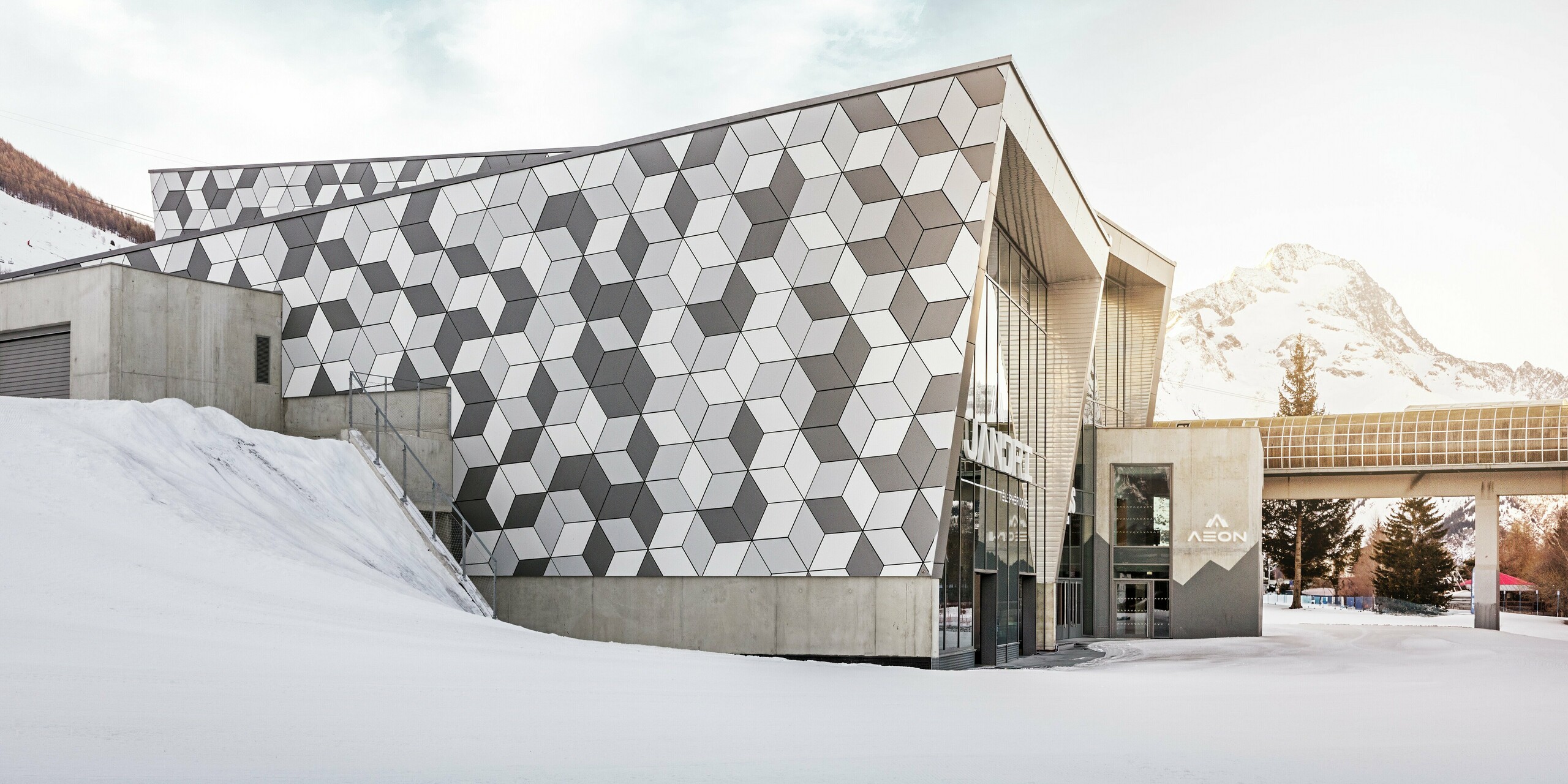 La station aval des Deux Alpes impressionne par sa façade exceptionnelle en panneaux composites en aluminium PREFABOND au motif losanges moderne. L'association de tons blancs et gris crée un design saisissant qui s'harmonise parfaitement avec le paysage montagneux enneigé. Cette façade durable et résistante aux intempéries garantit une solution esthétique et fonctionnelle, même dans des conditions extrêmes. Le soleil éclatant en arrière-plan crée un décor majestueux.